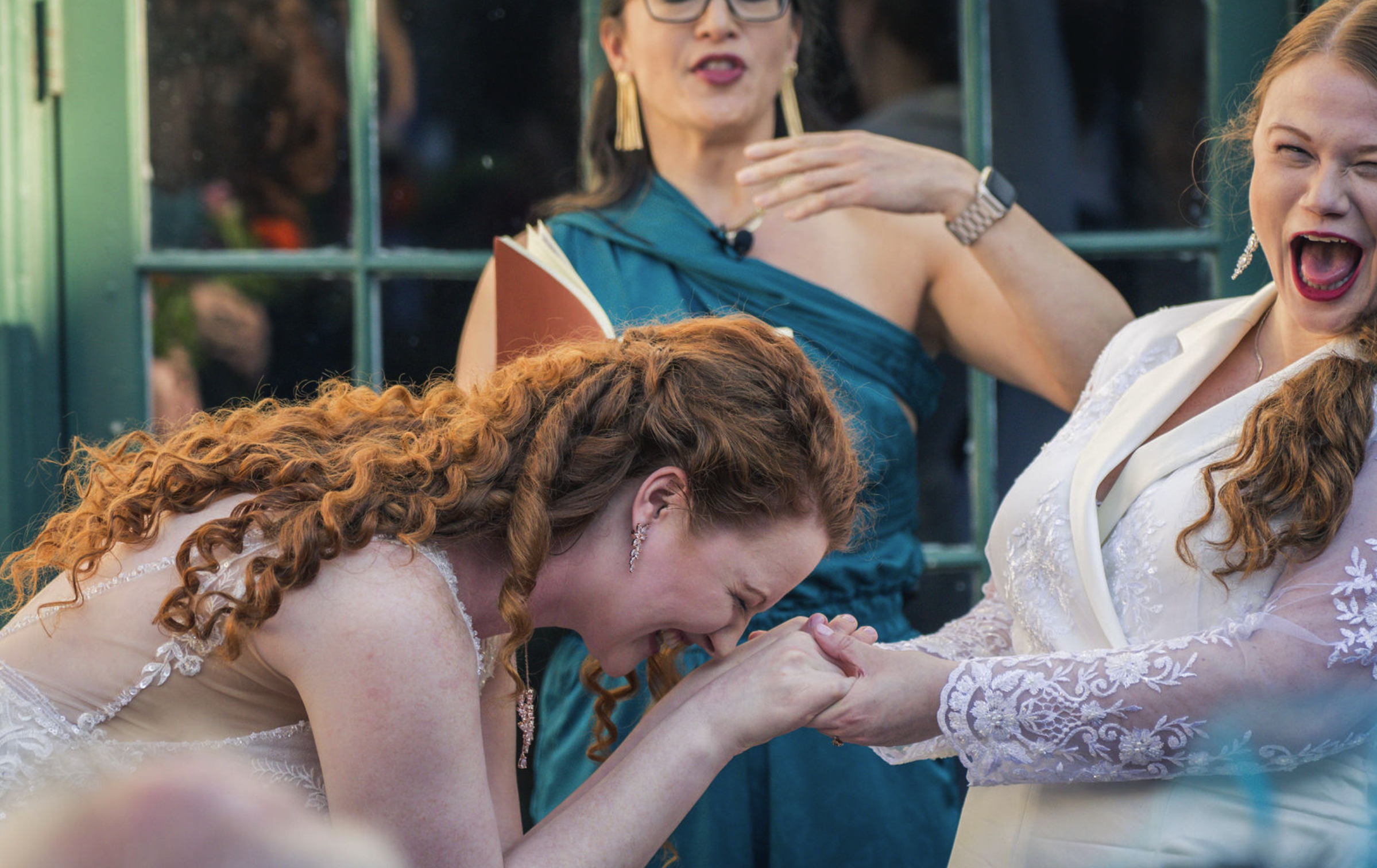 A woman with curly red hair in a wedding dress kisses the hand of another woman in a white lace dress, both smiling. Two women are behind them, one in teal dress with glasses, and the other in white laughing.