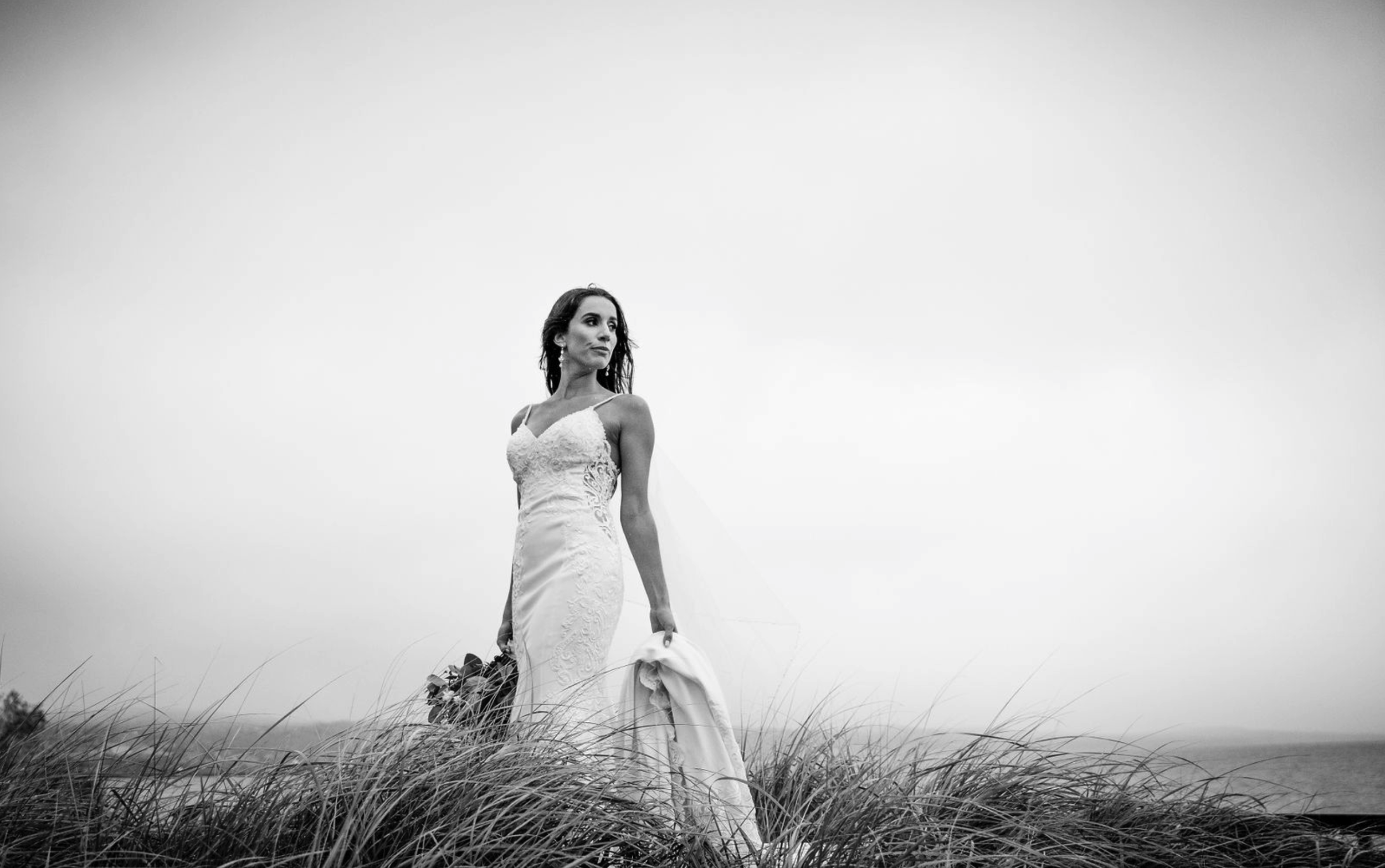 A woman in a wedding dress stands in tall grass near the coast, holding a bouquet, gazing to the side, in black and white.
