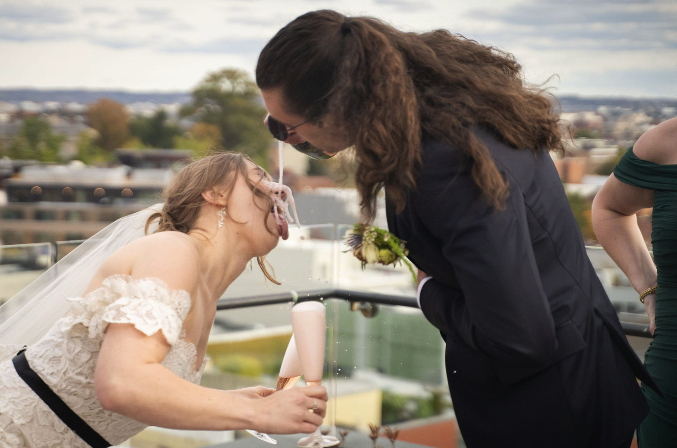 A bride and groom participate in a wedding tradition where the bride licks a spoon and the groom leans in for a kiss on a rooftop, with a cityscape in the background.