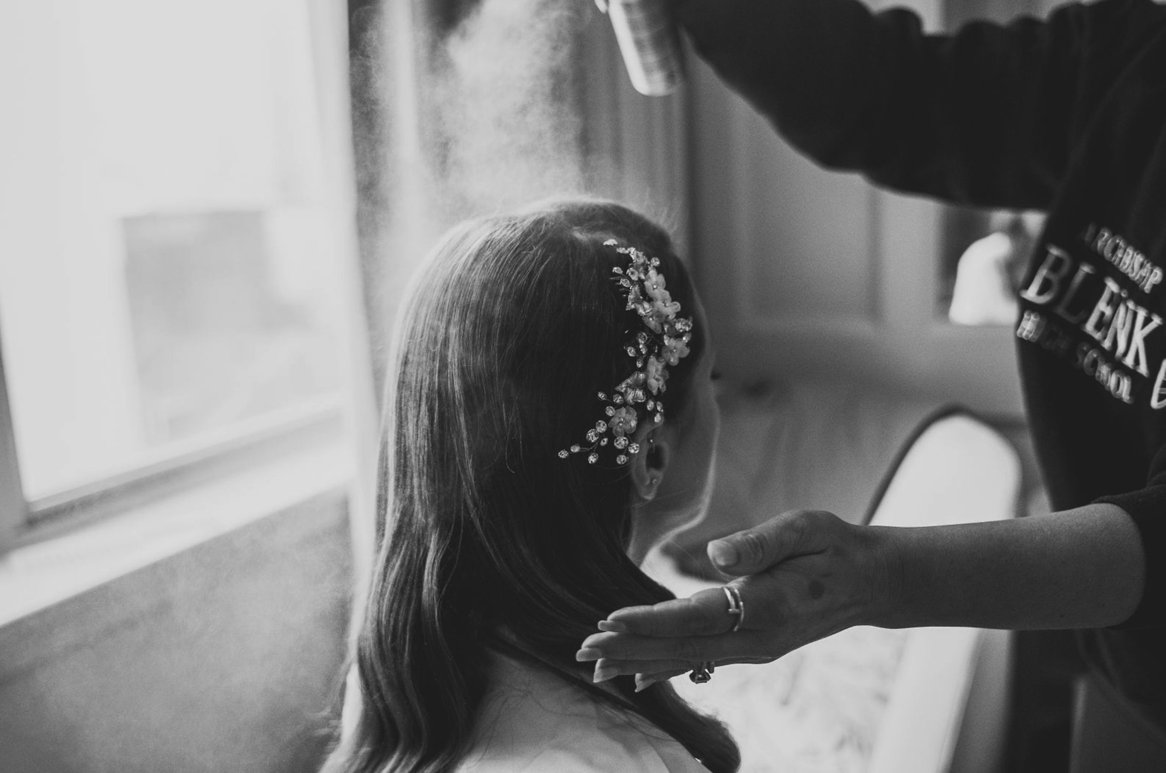 A woman with long hair, wearing a floral hair accessory, is having her makeup or hair done by another person in a room with natural light.