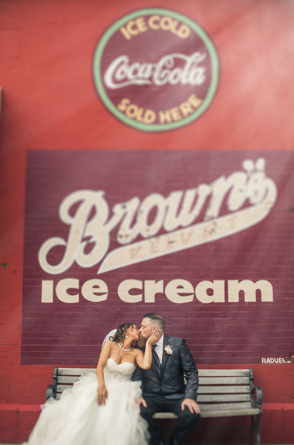 A newly married couple in wedding attire sitting on a bench, sharing a kiss in front of a large wall painting that says 'Brown's Ice Cream' and website information.