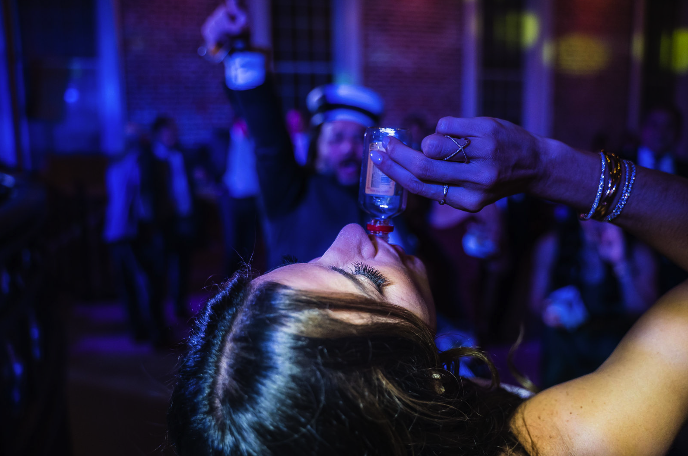 A woman at a party is lying on her back, drinking from a small bottle held by another person. The woman's eyes are closed, she has long eyelashes, and her hair is dark. The background shows a group of people in a dimly lit room with colorful lighting