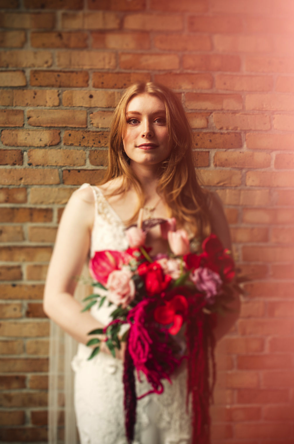 A New Orleans Pharmacy Museum wedding, featuring a beautiful young bride with long red hair holding a bouquet of pink and red flowers, standing against a brick wall, taken by J. Bellocq of Bellocq Weddings.