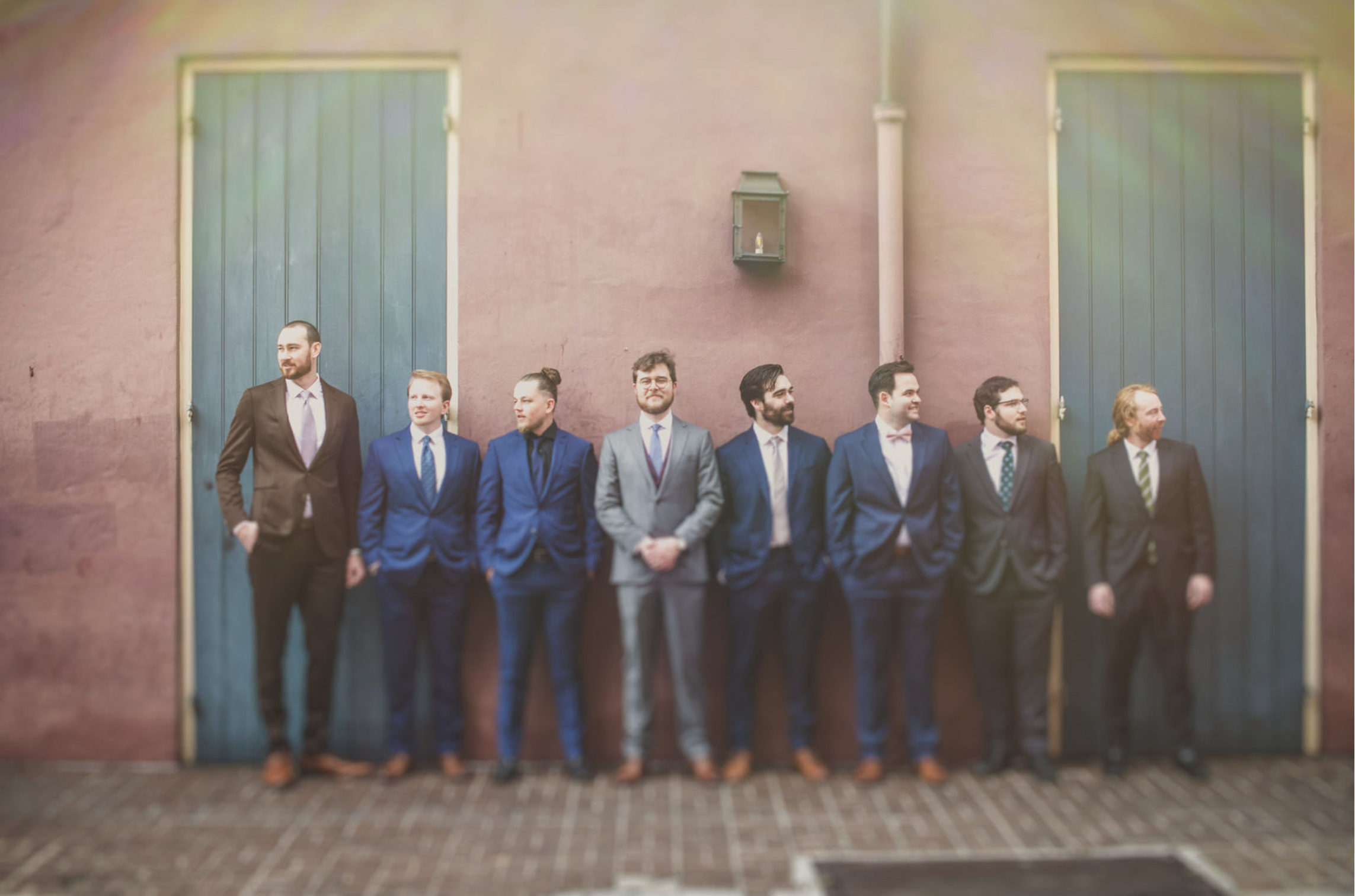 A group of eight groomsmen in formal suits standing against a pink wall with blue doors, posing for a photo, taken by J. Bellocq, luxury wedding photojournalist of Bellocq Weddings in the French Quarter of New Orleans.