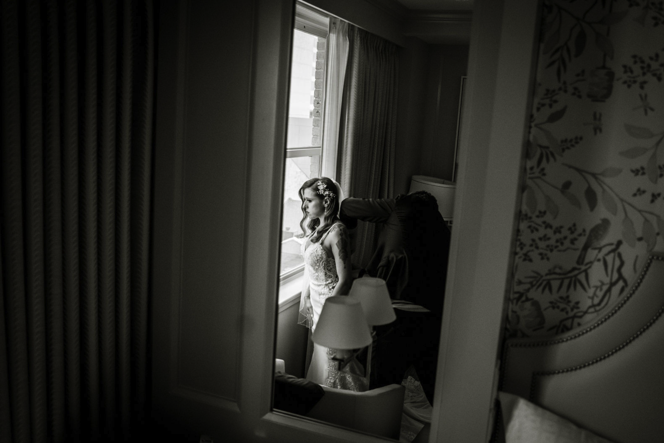 A bride getting ready, standing by a window with natural light, her hair styled with a floral accessory, as someone helps her with her dress.