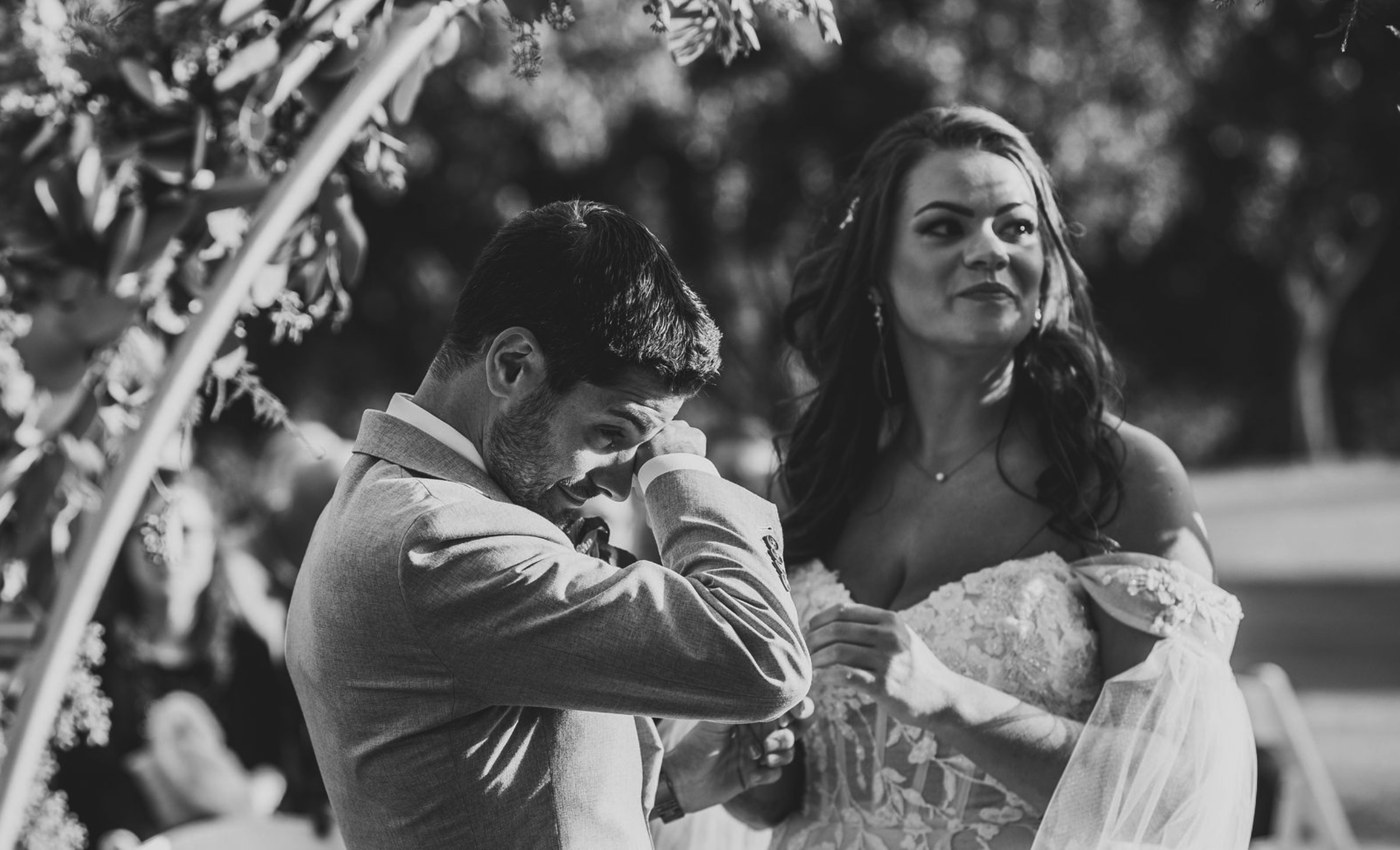 A black-and-white photo of a man in a suit crying and covering his face with his arm, standing next to a woman in a wedding dress with wavy hair, outdoors with blurred trees in the background, during a wedding ceremony.