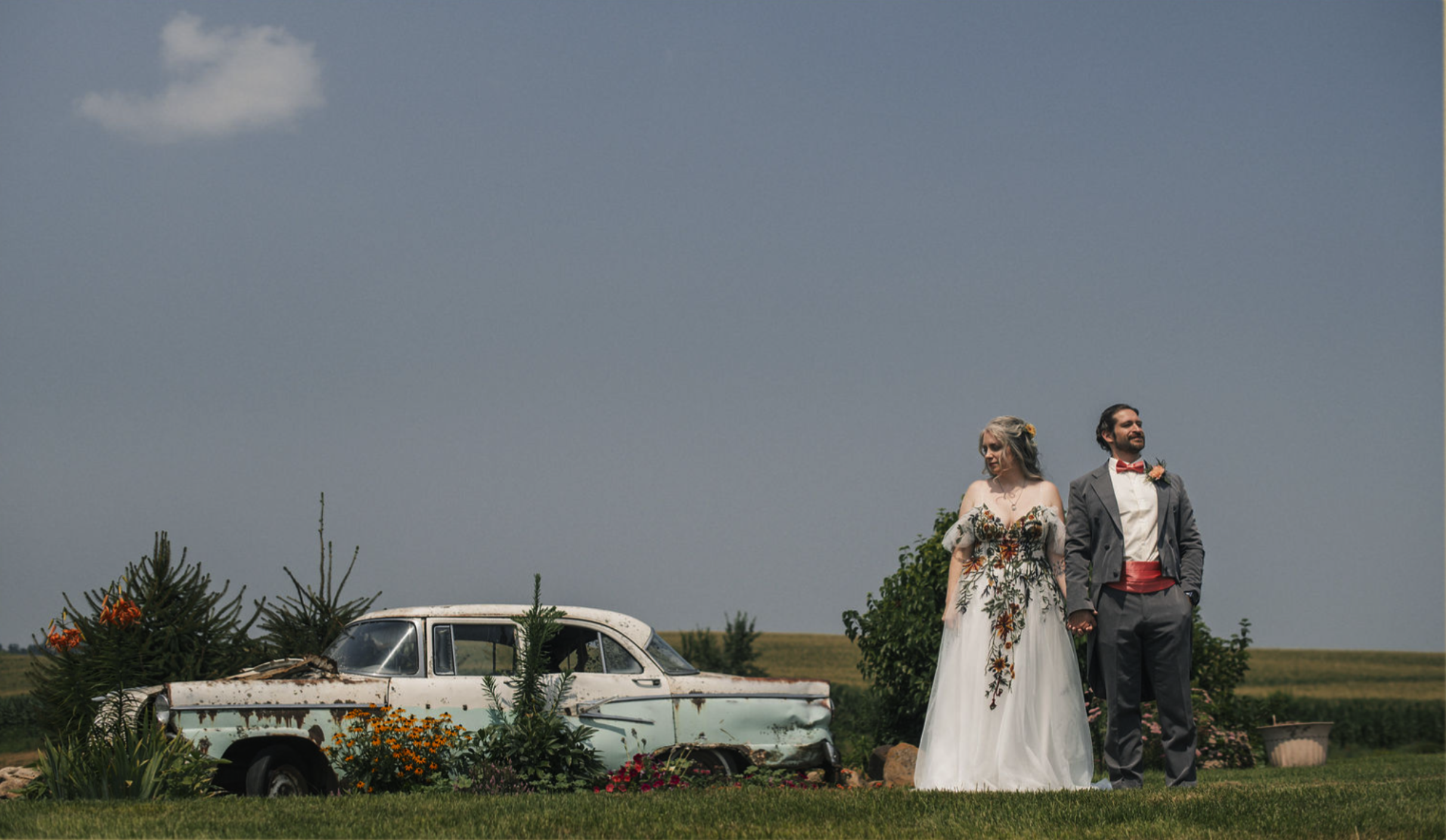 A newlywed couple holding hands stands outdoors in front of an old rusty car surrounded by flowers and greenery under a blue sky with clouds, taken by J. Bellocq, luxury wedding photojournalist of Bellocq Weddings taken on a farm in Minnesota.