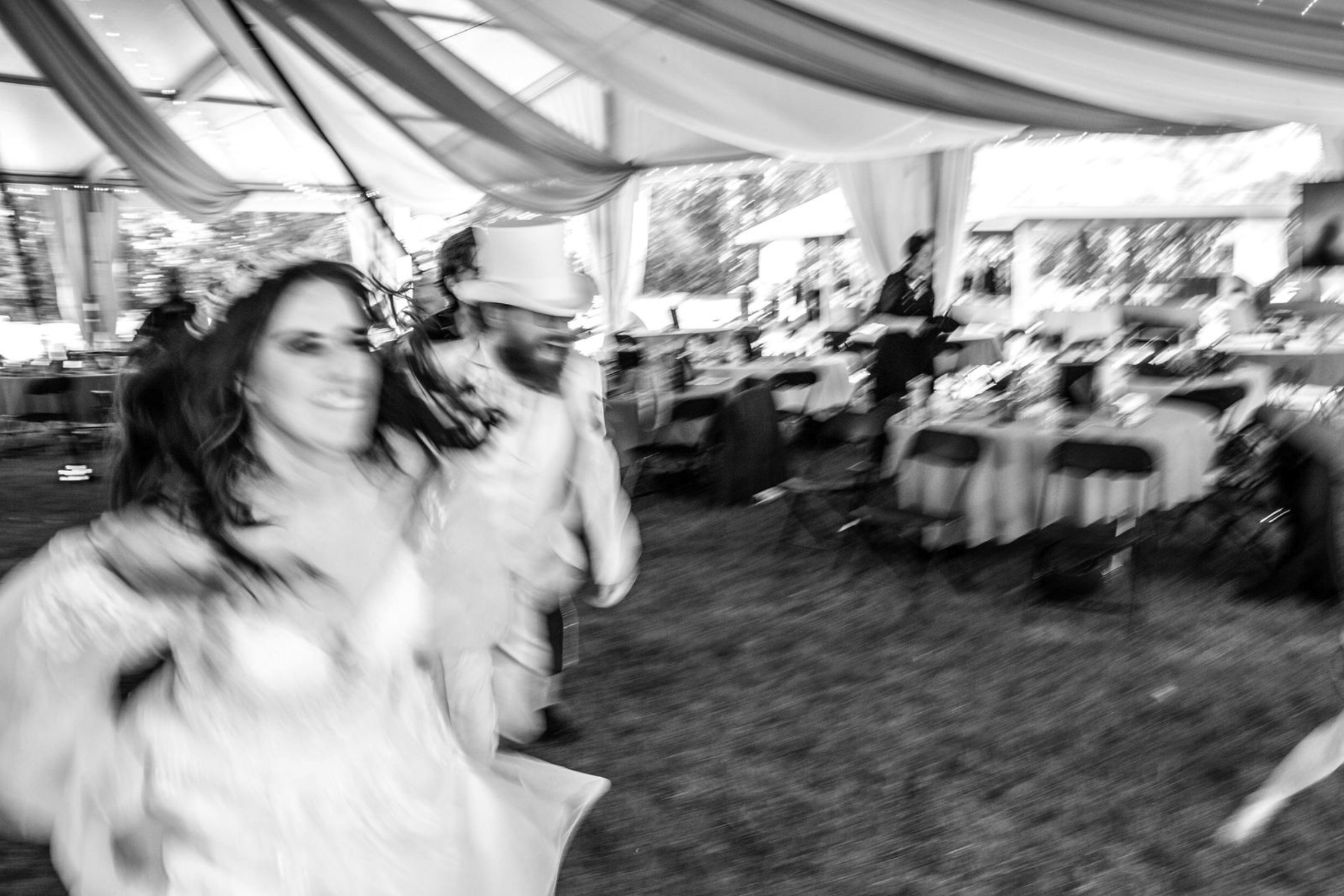 Blurry black and white photo of a woman and a man in a tented event space, with tables and chairs in the background. The woman is smiling, and the man wearing a hat is next to her.
