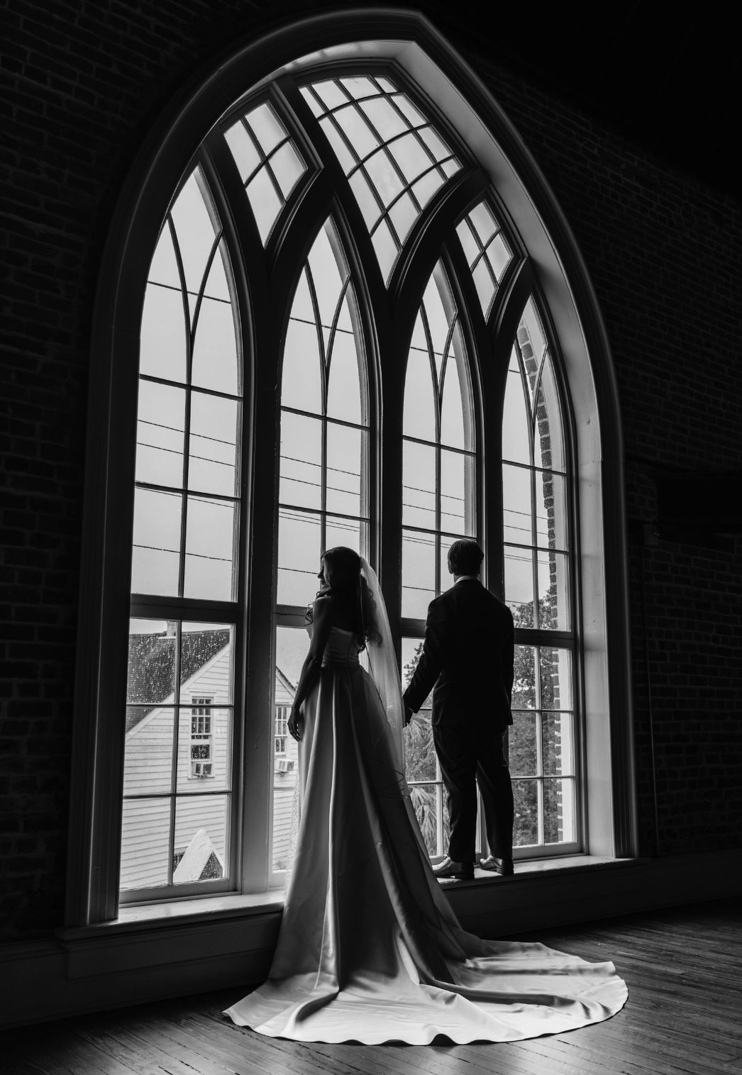 Felicity, New Orleans. A luxury black and white photo of a bride and groom standing by a large arched window, holding hands and looking out, taken by J. Bellocq of Bellocq Weddings.