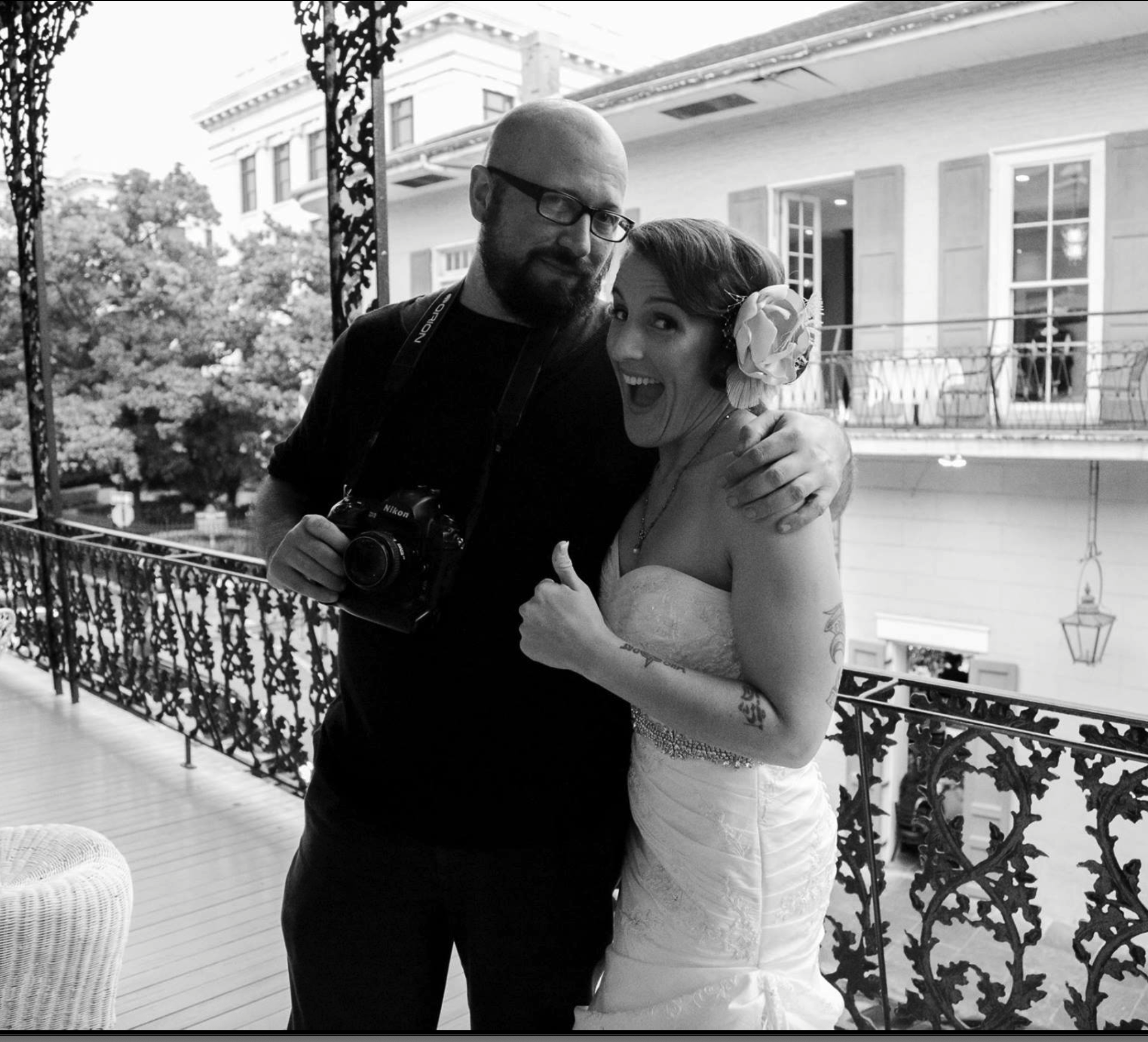 A man with a camera and a woman in a wedding dress are hugging on a balcony with ornate railing. The woman has a large flower in her hair and tattoos on her arm, and is giving a thumbs-up.