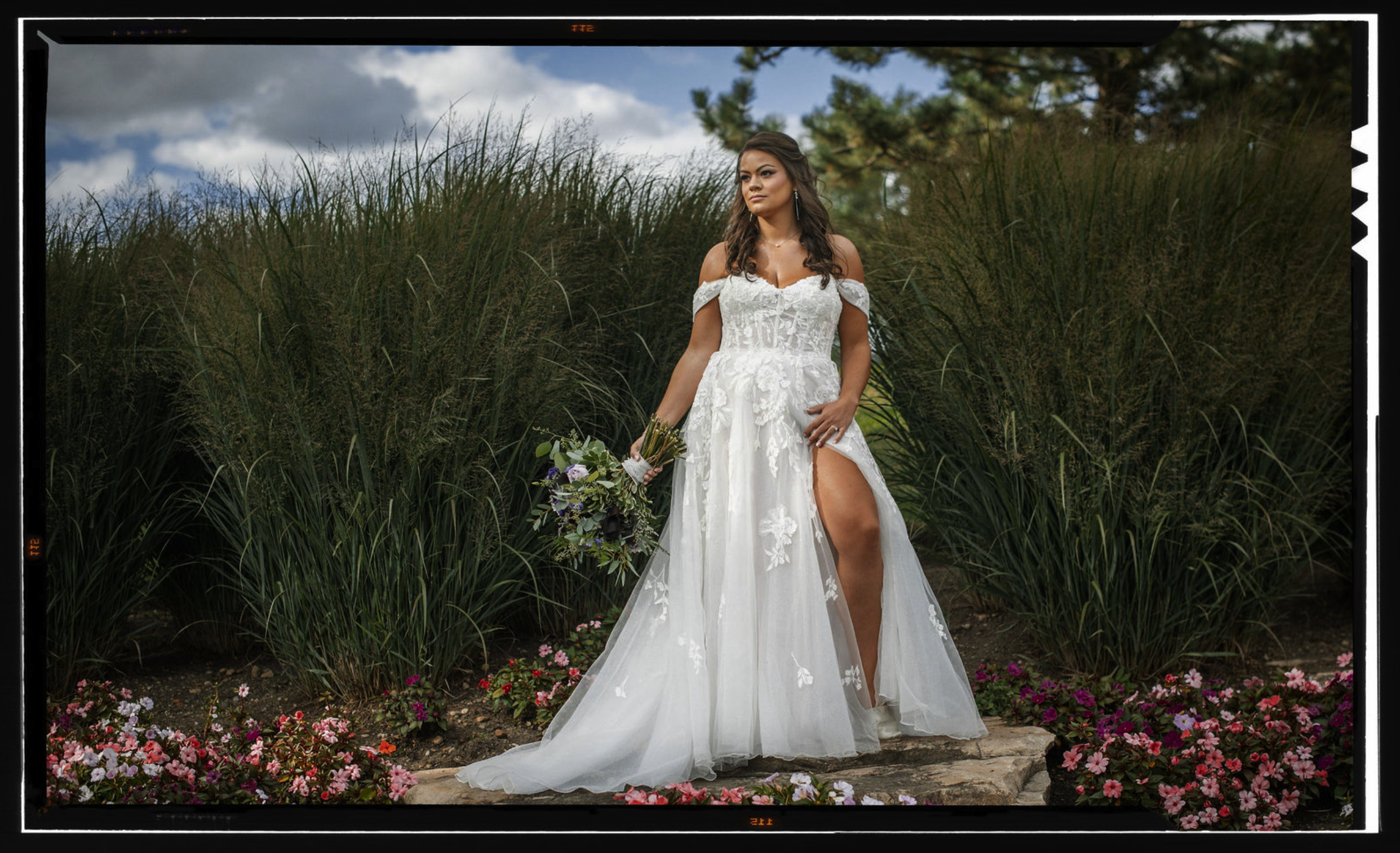 A woman in a white wedding gown with lace details, holding a bouquet of flowers, standing outdoors on a stone path surrounded by tall grasses and colorful flowers, with a cloudy sky in the background.
