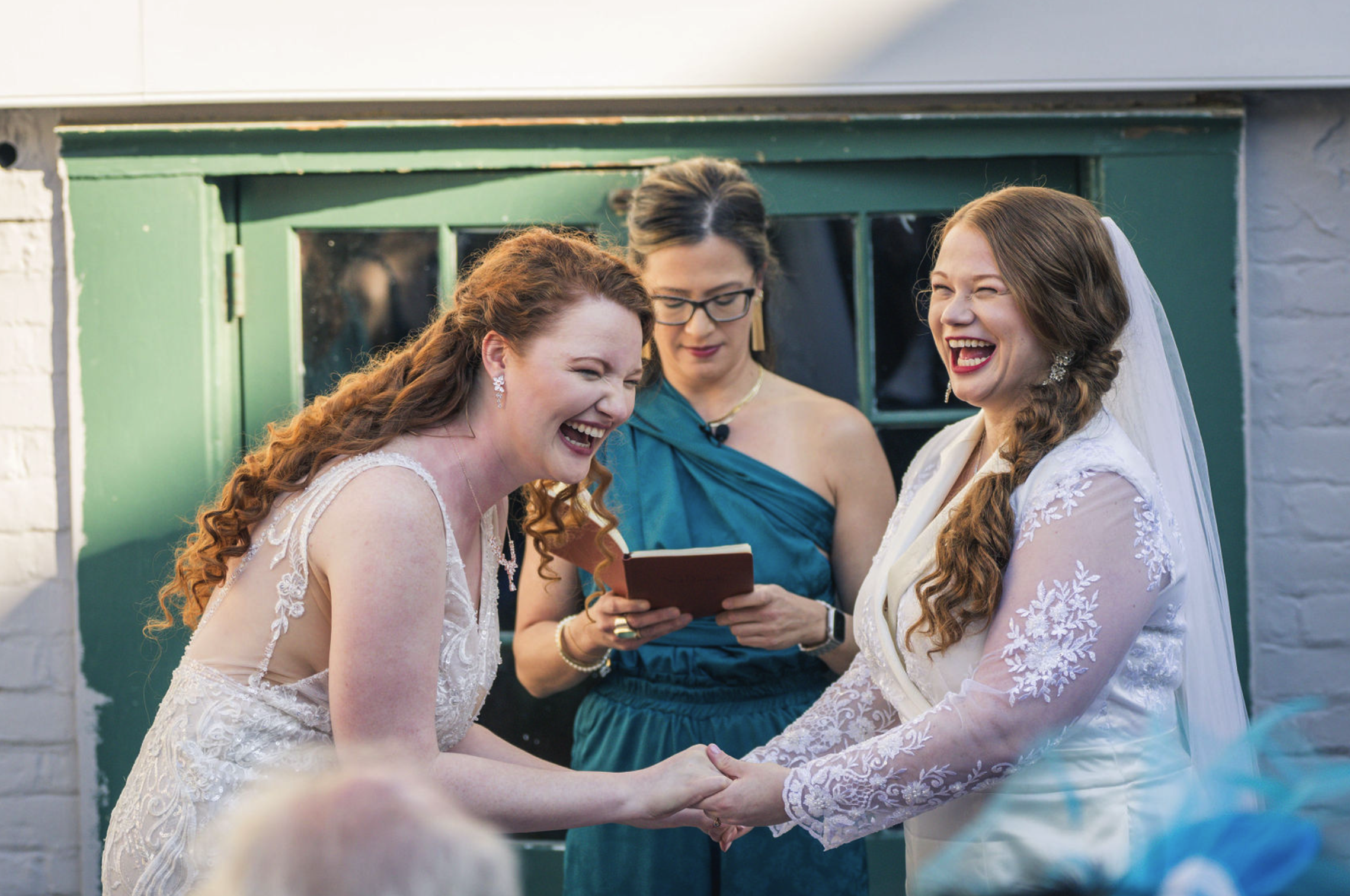 Two women, a bride and a bride, holding hands and smiling during a wedding ceremony, with a woman in a teal dress officiating in the background.