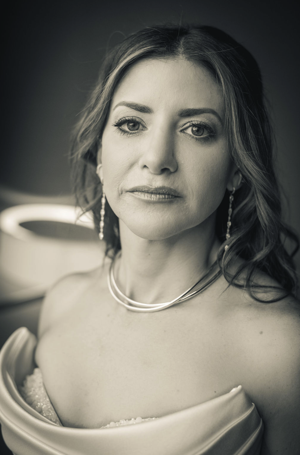 A luxury Felicity New Orleans black-and-white portrait of a bride with shoulder-length wavy hair, wearing earrings and layered necklaces, gazing directly at the camera with a serious expression, taken by J. Bellocq of Bellocq Weddings.