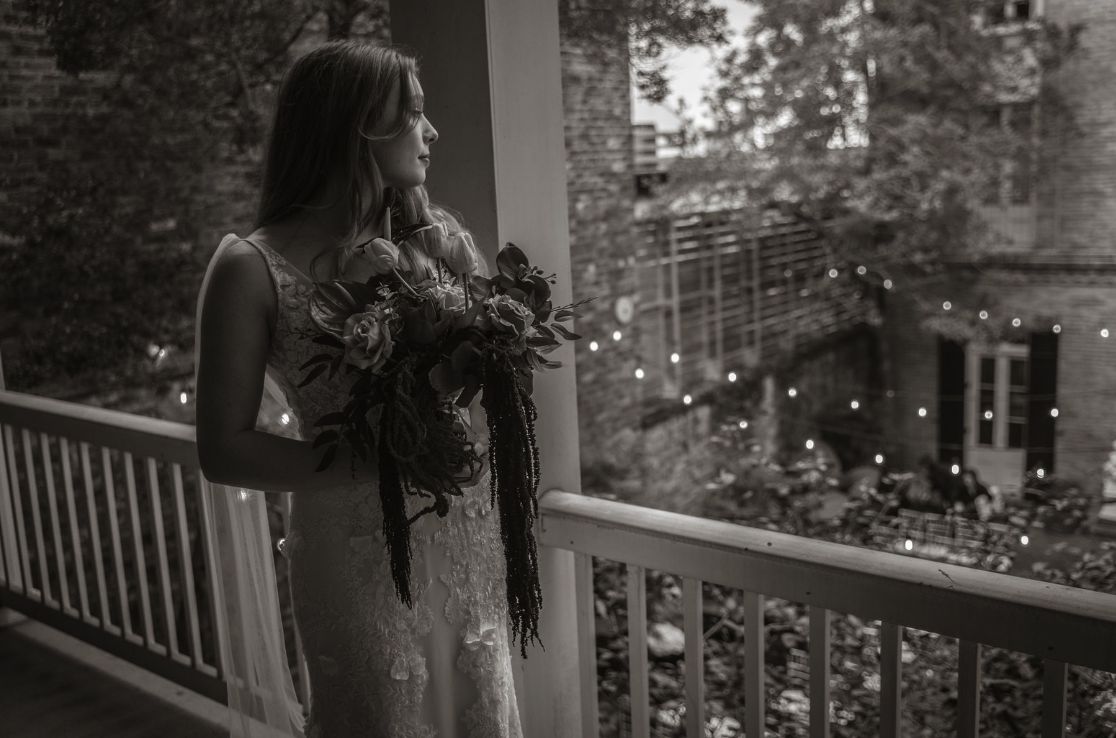 A woman in a wedding dress holding a bouquet, standing on a balcony and looking outside at a blurred urban scene with string lights, in black and white.
