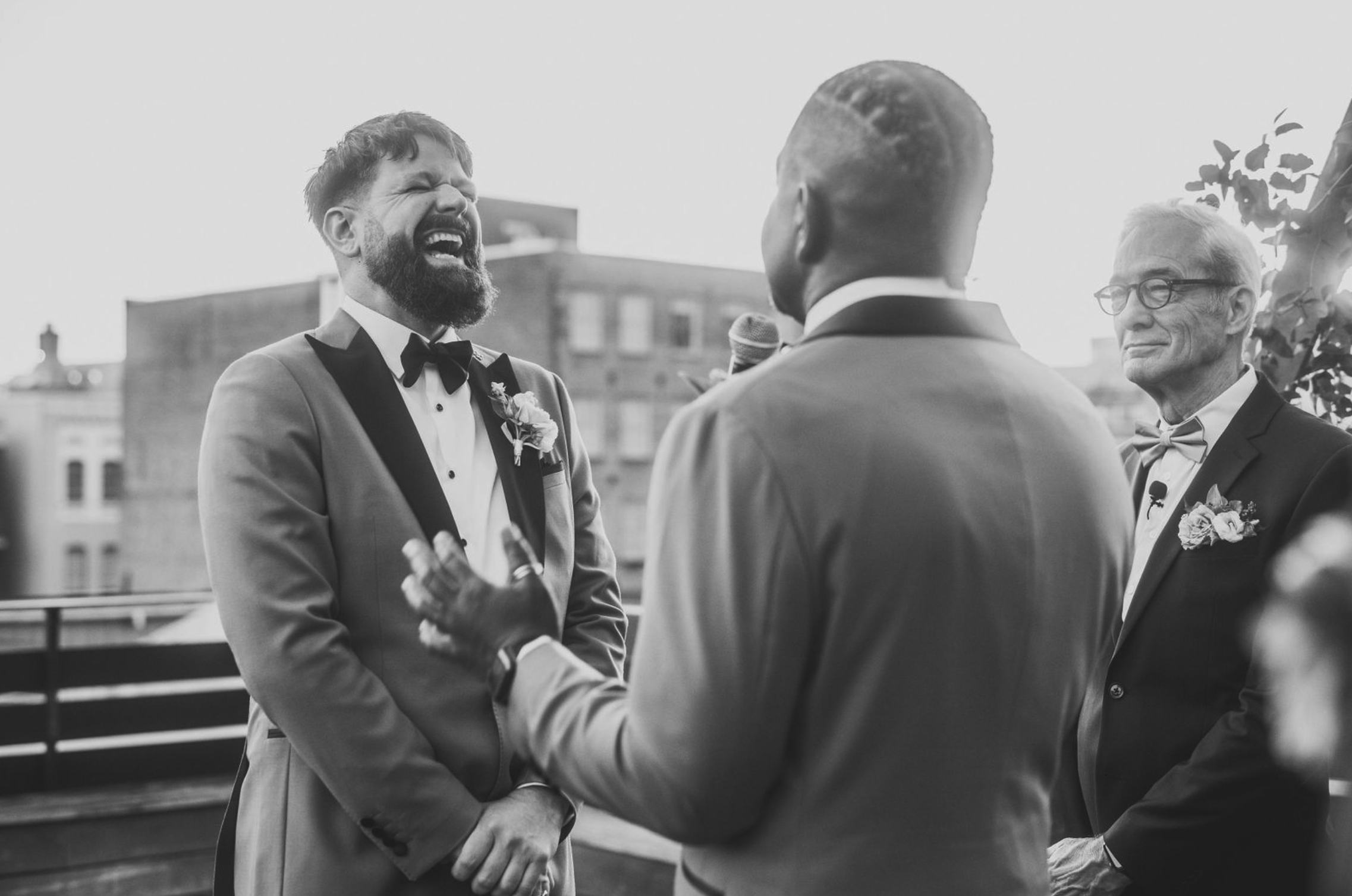 Two men in tuxedos, one with a beard singing or speaking emotionally, during a wedding ceremony on a rooftop, with another man holding a microphone and an older man with glasses smiling in the background.