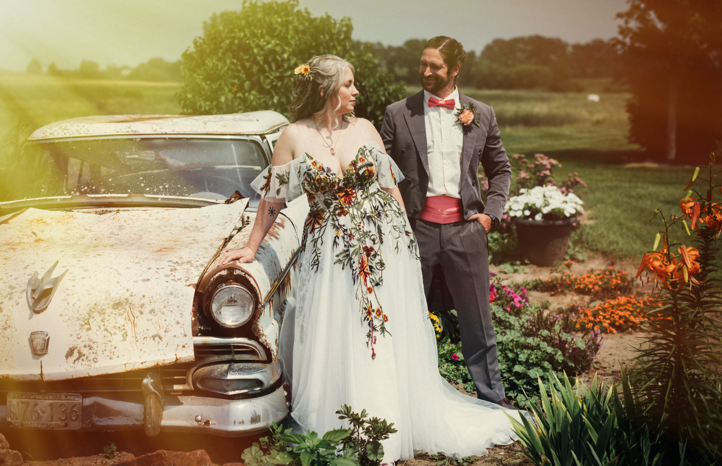 A woman in a white floral dress and a man in a suit with a pink bowtie pose next to a rusty, vintage car in a garden with flowers and greenery, taken by J. Bellocq, luxury wedding photojournalist of Bellocq Weddings on a beautiful farm in Minnesota.