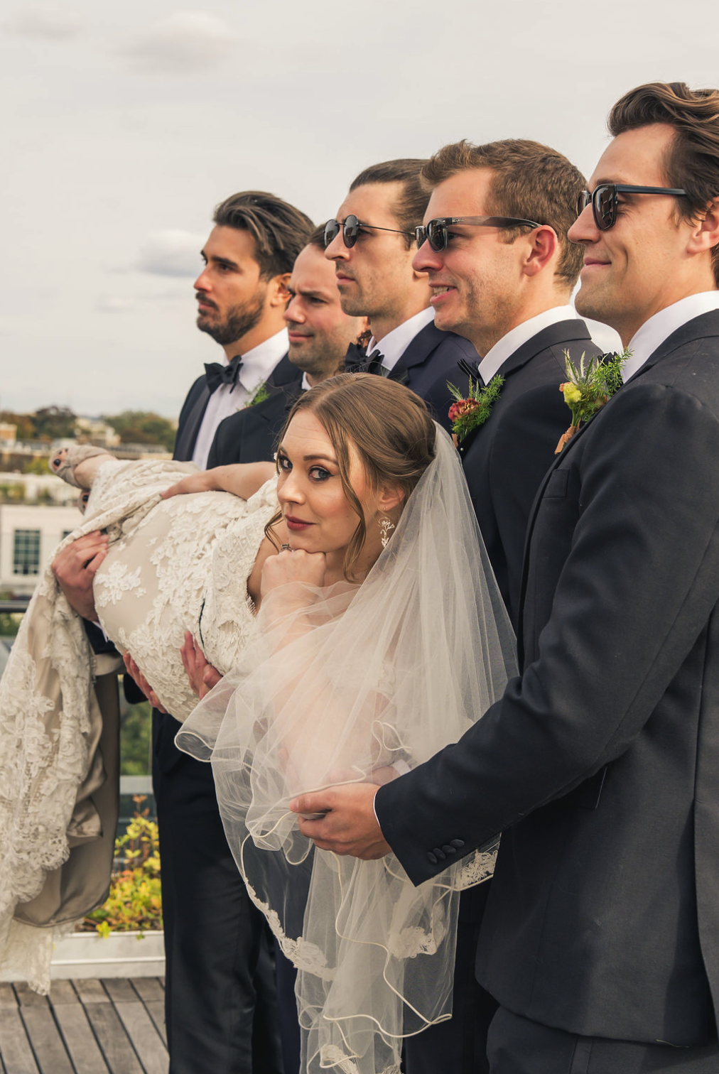 A bride being carried on the shoulder of a groomsman at her wedding, with five men in tuxedos and sunglasses standing behind her, on an outdoor terrace with a cityscape in the background.