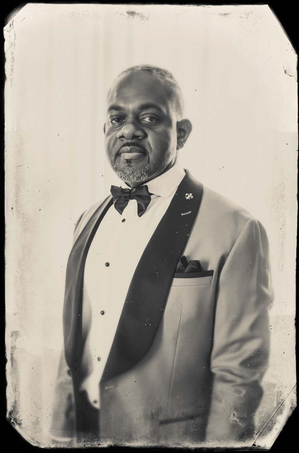 A stunning African-American groom in formal tuxedo with a bow tie, pocket square, and a lapel pin, posing for a black-and-white portrait, taken by J. Bellocq, luxury wedding photojournalist of Bellocq Weddings in New Orleans.