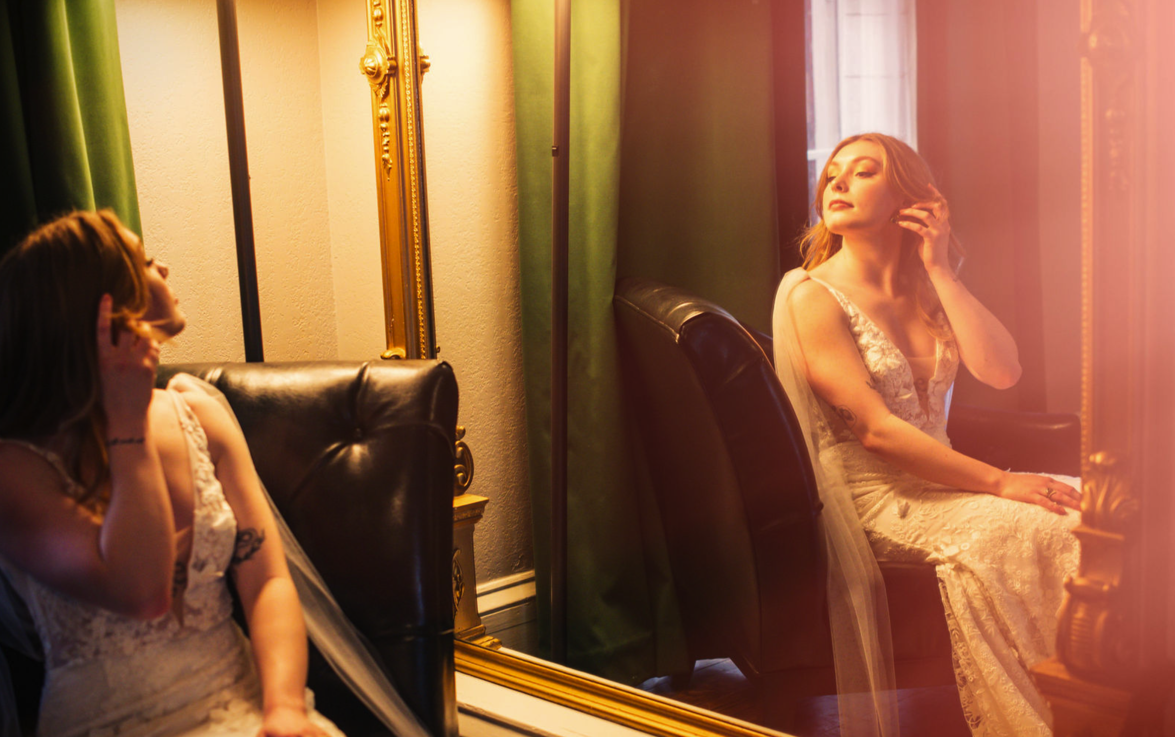 Woman in a lace dress sitting in front of a mirror, adjusting her hair, as she looks at her reflection, with green curtains and a gold-framed mirror behind her.