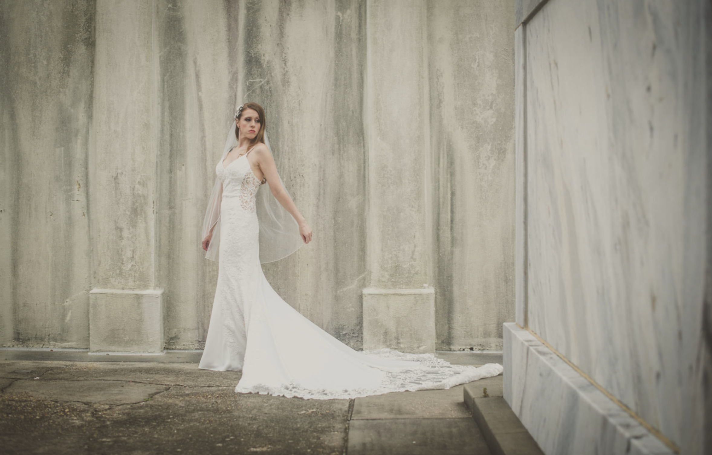 A woman in a white wedding dress with a long train standing against a concrete wall, looking to her left.