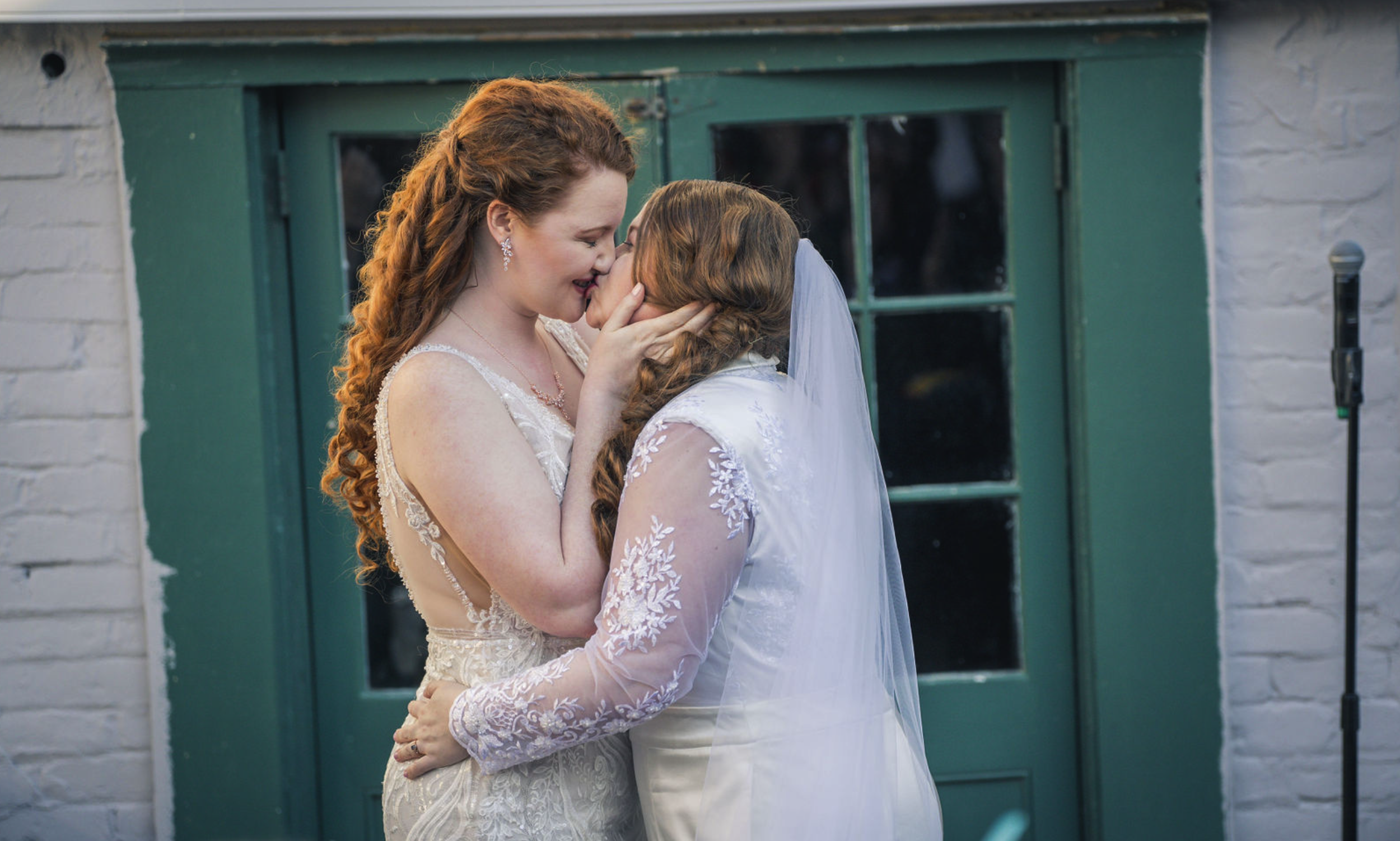 Two women in wedding dresses sharing a kiss, with one cradling the other's face, in front of a green door and white brick wall. One woman has long curly red hair, the other has long straight hair with a veil.