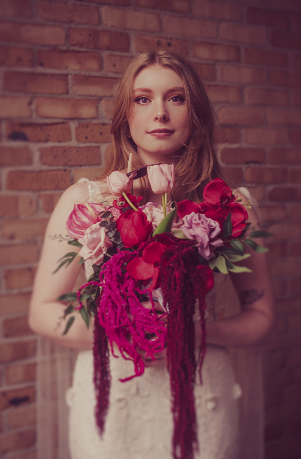 A woman with long, wavy hair wearing a white dress, holding a vibrant bouquet of pink, red, and purple flowers in front of a brick wall, taken by J. Bellocq, luxury wedding photojournalist of Bellocq Weddings in New Orleans.