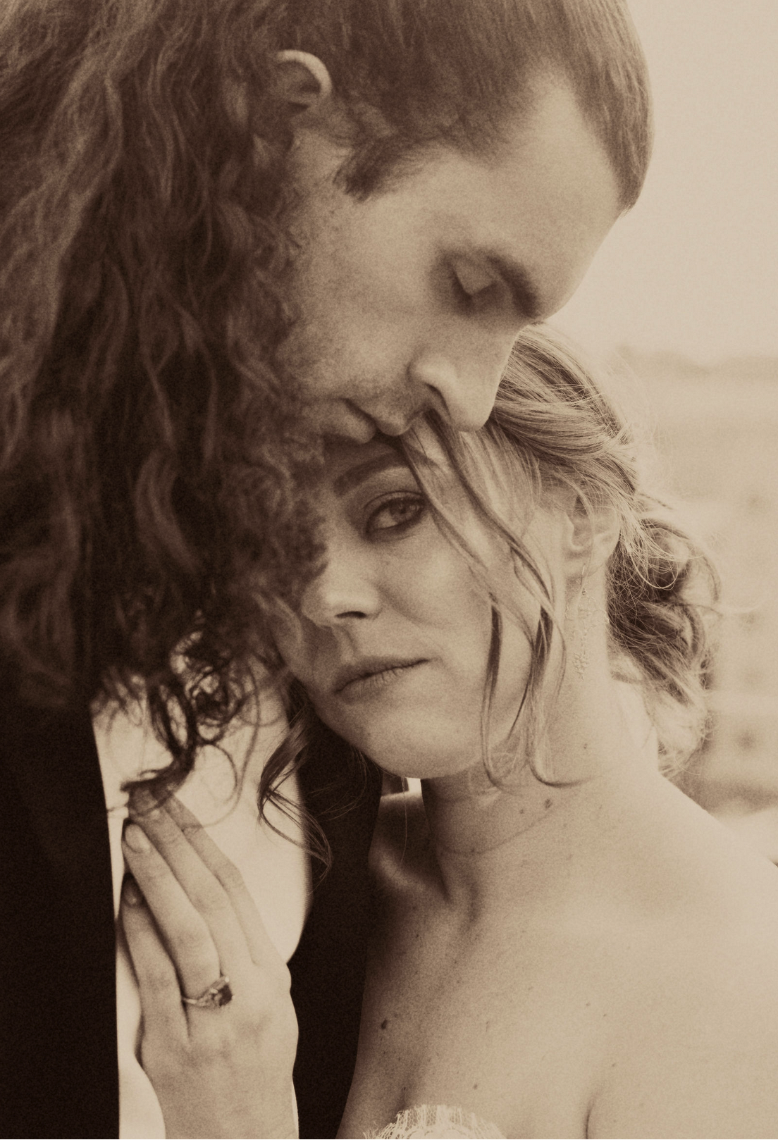 A close-up editorial sepia photo of a bride and groom, with the man gently resting his forehead on the woman's. The woman looks directly at the camera with a serious expression, taken by J. Bellocq, luxury wedding photojournalist of Bellocq Weddings