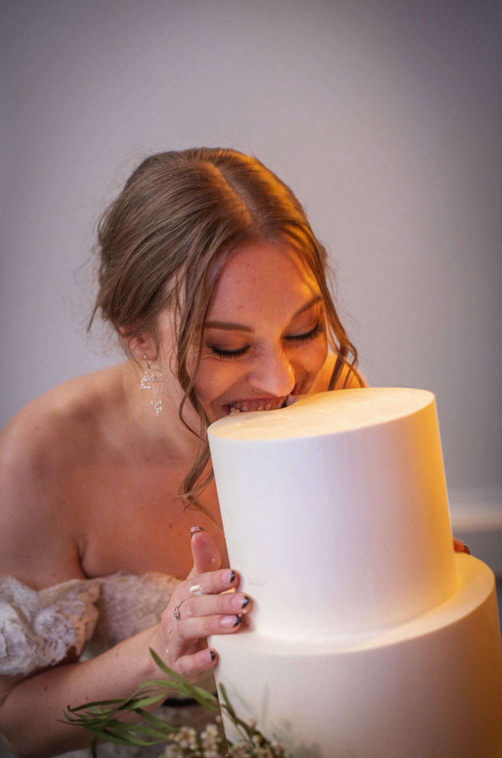 A bride in a wedding dress is smiling and pretending to bite into a tall, white wedding cake.