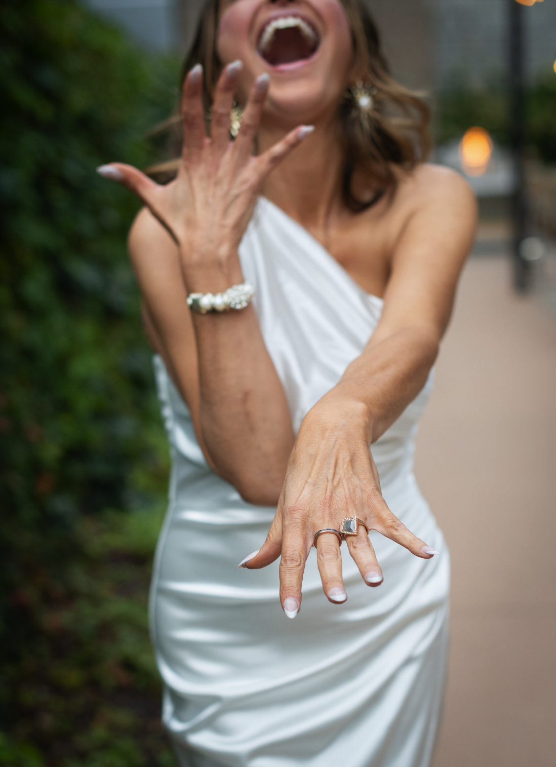 A woman in a white dress showing off her wedding rings and jewelry, laughing with her mouth open.