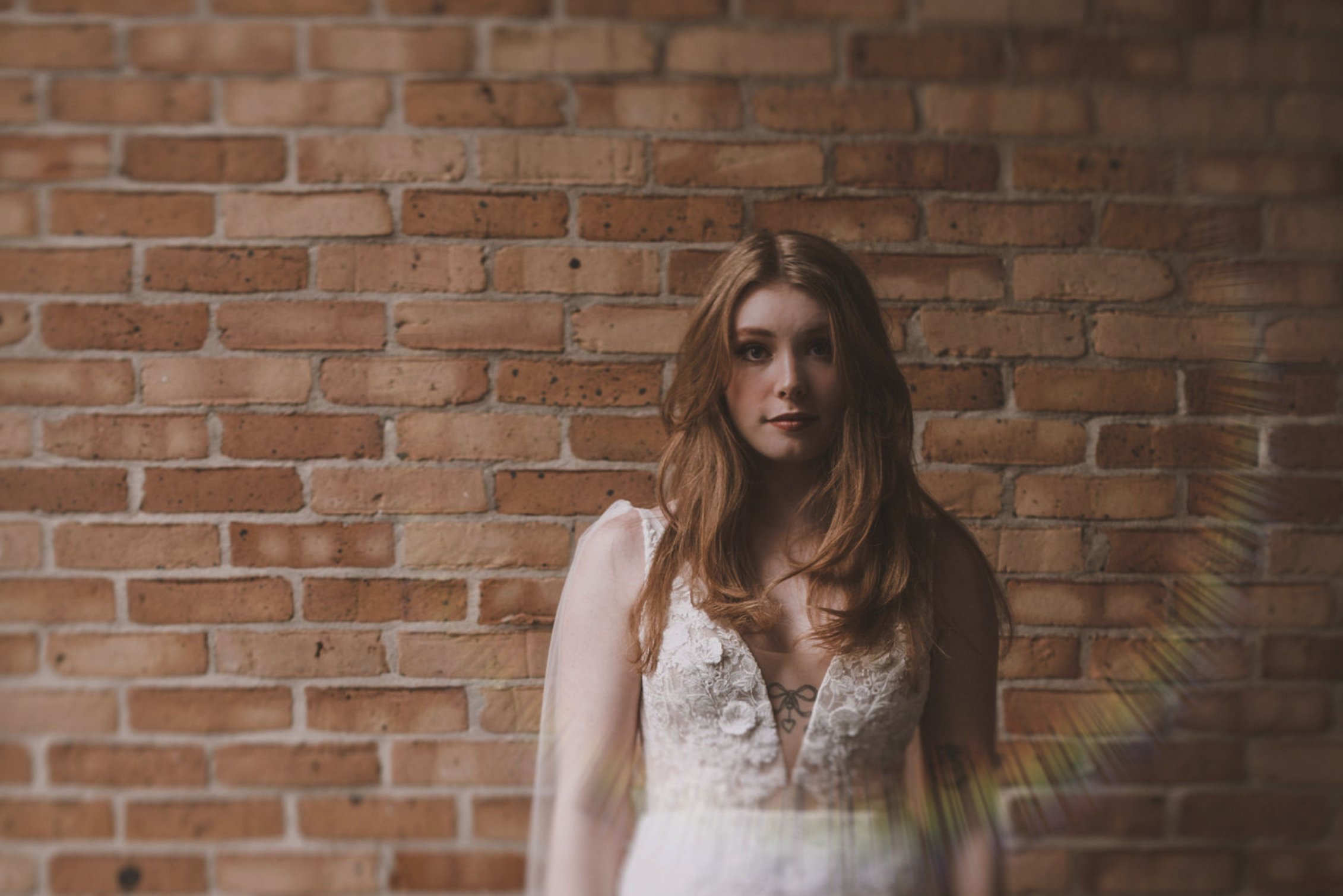 A young woman with wavy auburn hair and fair skin standing against a brick wall, wearing a white dress with lace details and a small tattoo visible on her chest.