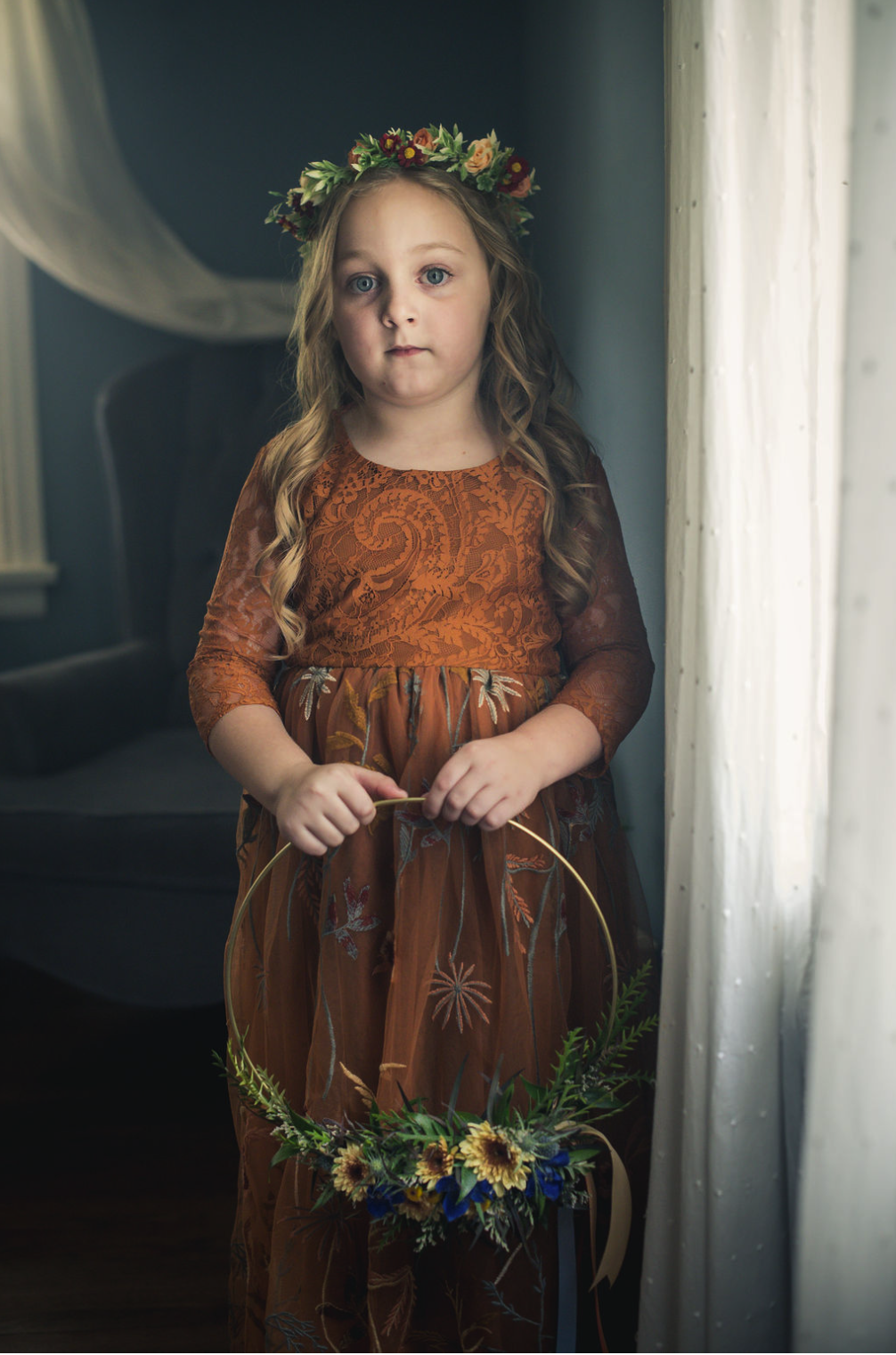 A young flower girl with long blonde curly hair, wearing a brown floral lace dress and a flower crown, stands by a window holding a sunflowers and greenery wreath, taken by J. Bellocq of Bellocq Weddings.
