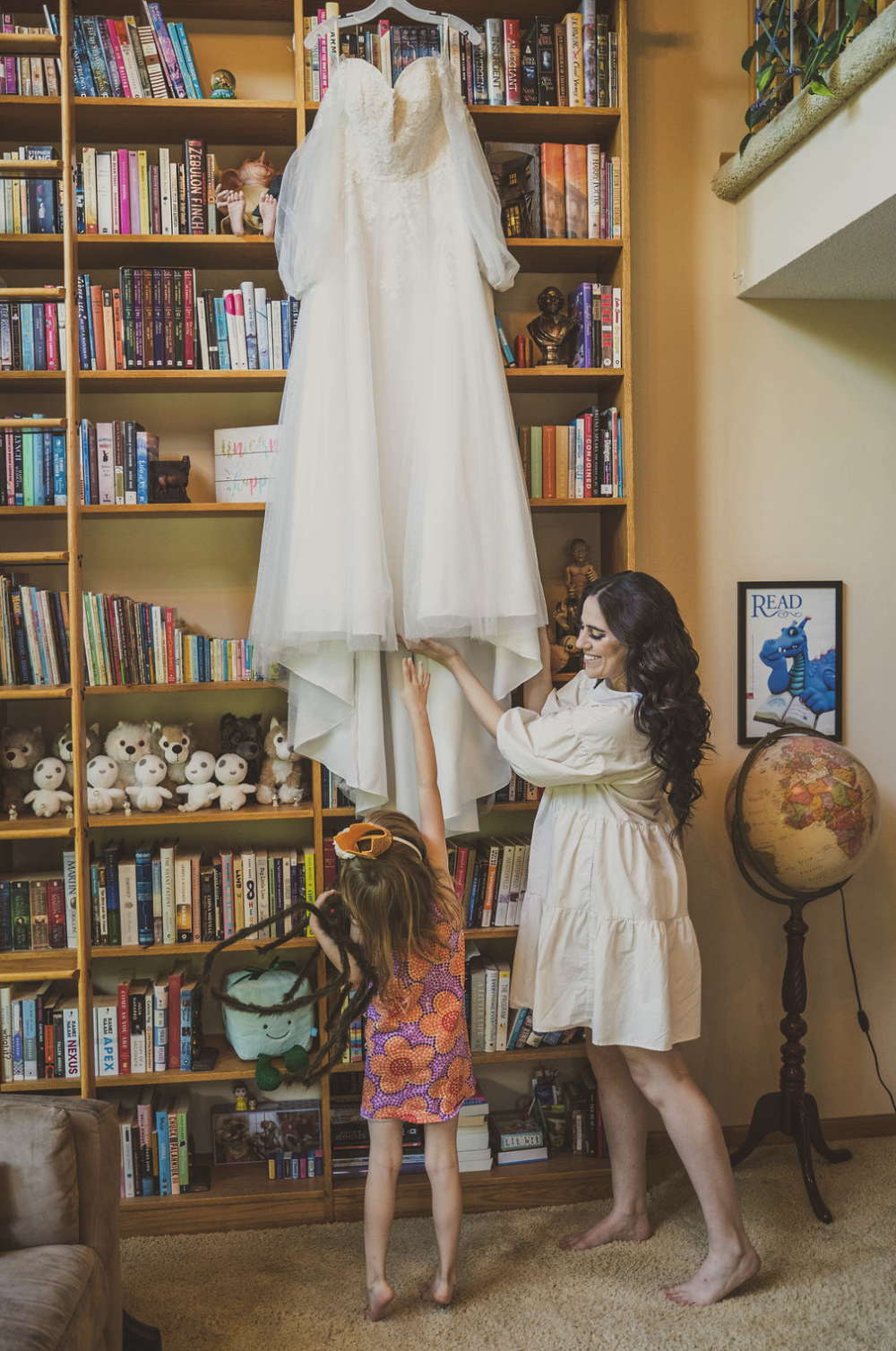 A woman and a young girl are hanging a white wedding dress on a bookshelf in a cozy room. The woman is smiling, and the girl, wearing a colorful dress with a bow in her hair, reaches up to touch the dress. The bookshelf is filled with books, stuffed 
