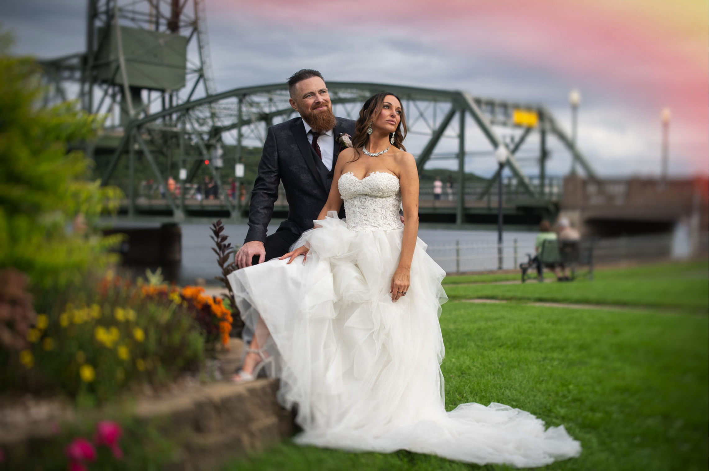 A bride and groom posing outdoors near a river with a bridge in the background on their wedding day. The bride is sitting on a ledge in a white wedding gown, and the groom is kneeling beside her in a dark suit. The sky is cloudy with a hint of pink s