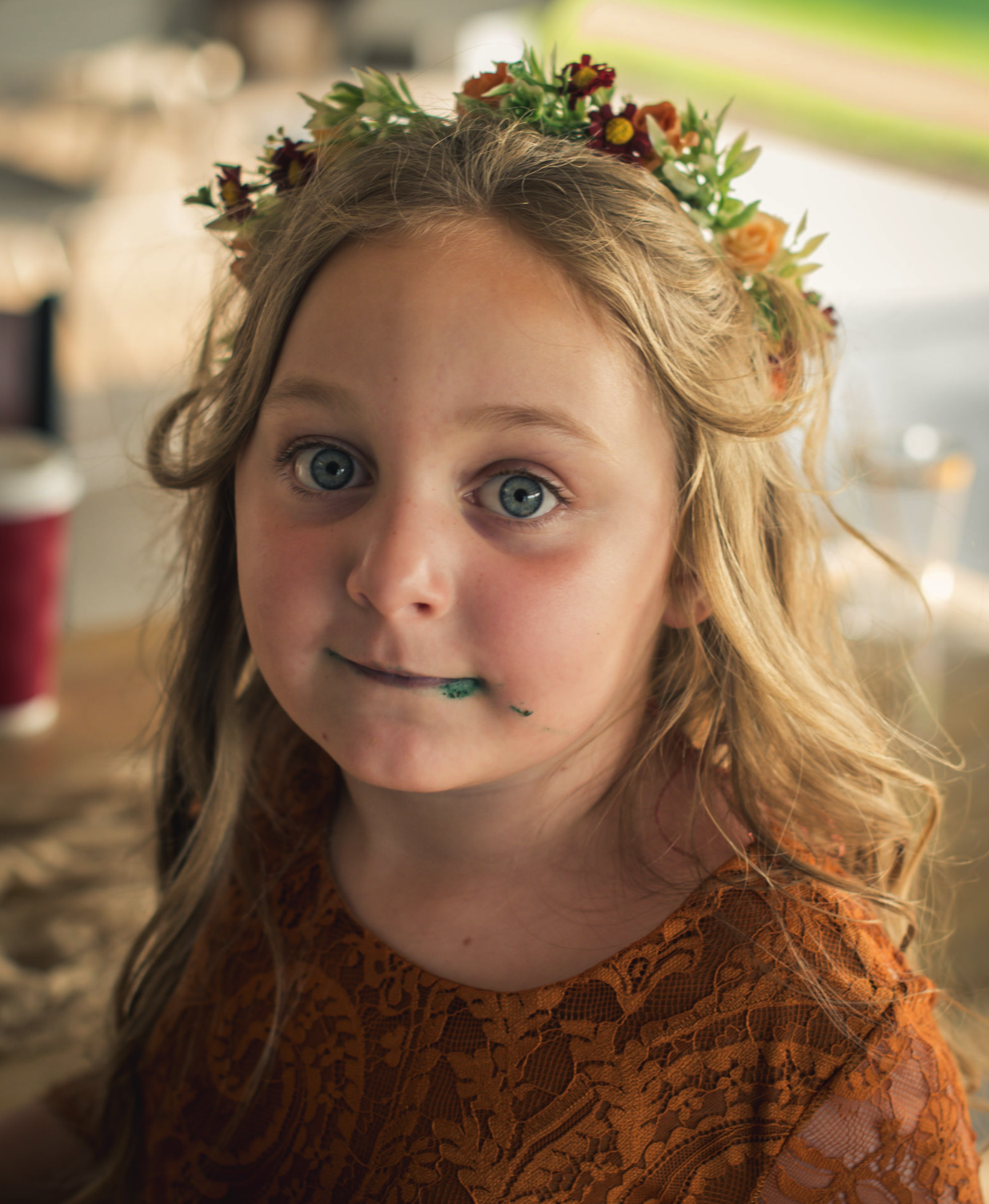 A young girl with blue eyes and curly blonde hair wearing a floral crown and an orange lace dress, with a small amount of green paint on her lips.