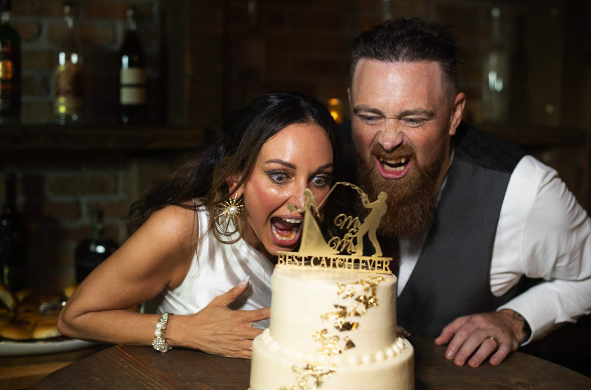 A couple celebrating a wedding or special event, leaning over a cake with a gold topper that reads 'Best Catch Ever' and depicts a fishing scene, in a cozy, rustic setting., taken by J. Bellocq, luxury wedding photojournalist of Bellocq Weddings in N