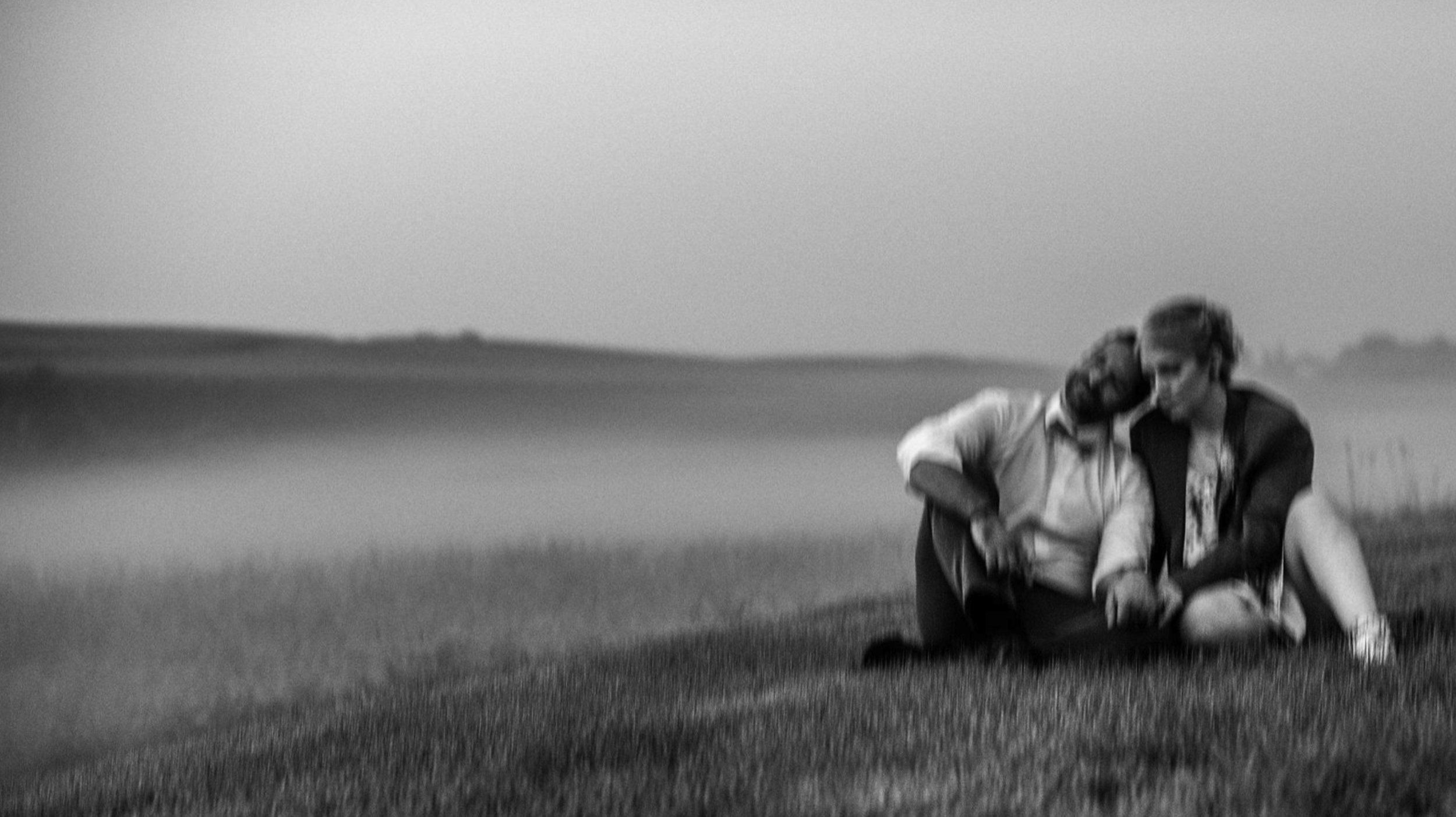 A black and white photo of a young couple sitting on grass in a field, sharing a tender moment with their heads leaning together.