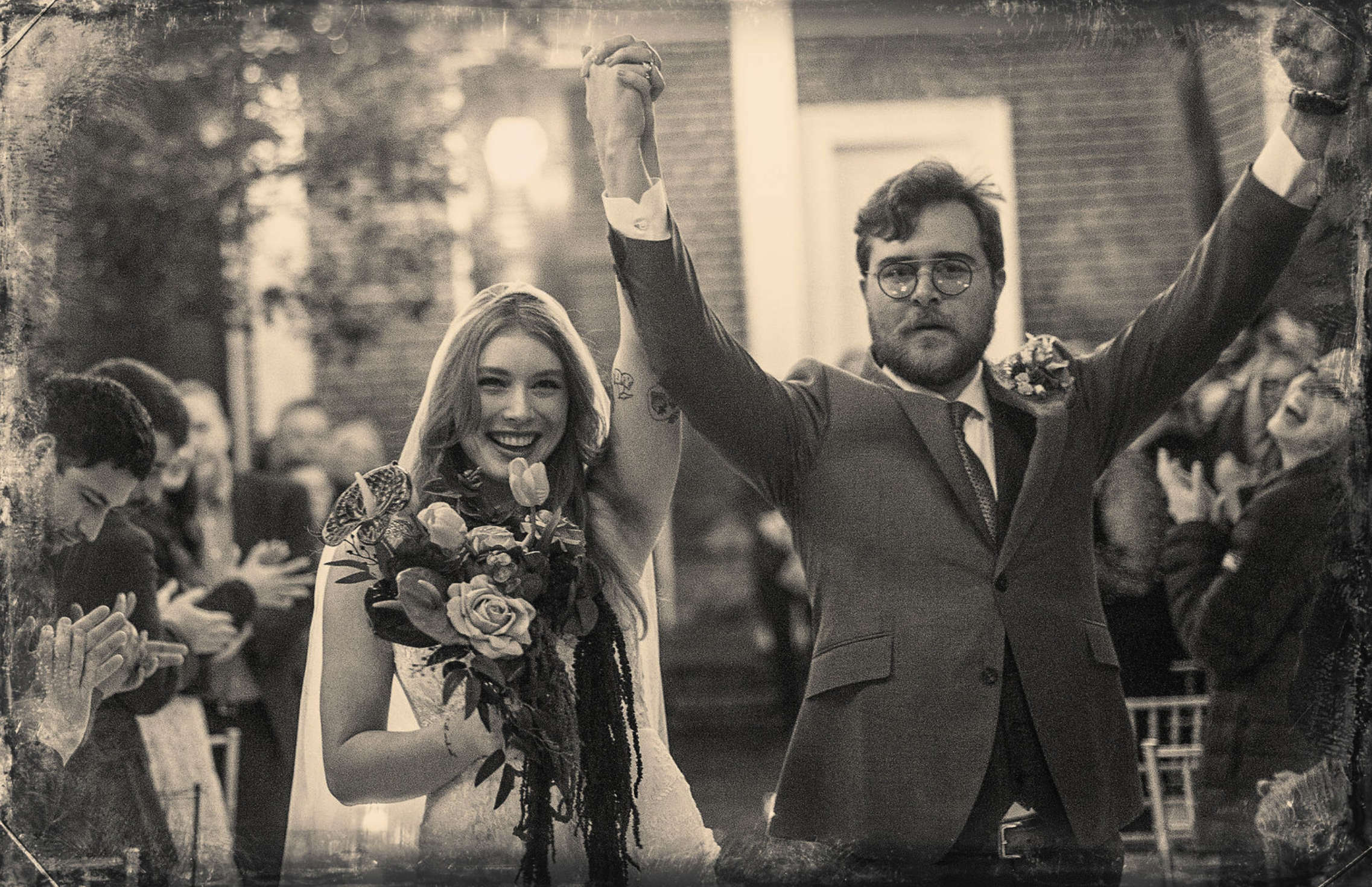 A newlywed couple celebrating, with the bride holding a bouquet of roses and the groom raising his arm in victory, during a wedding ceremony outdoor.