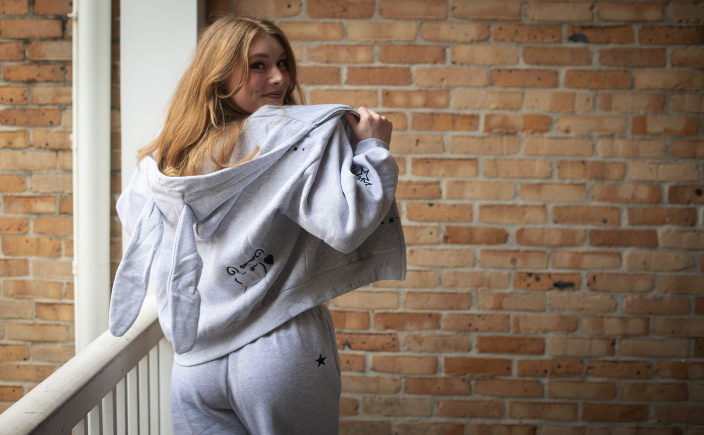A young woman with red hair standing on a balcony against a brick wall, wearing a gray sweatshirt and sweatpants, holding a gray hoodie with bunny ears over her shoulder.