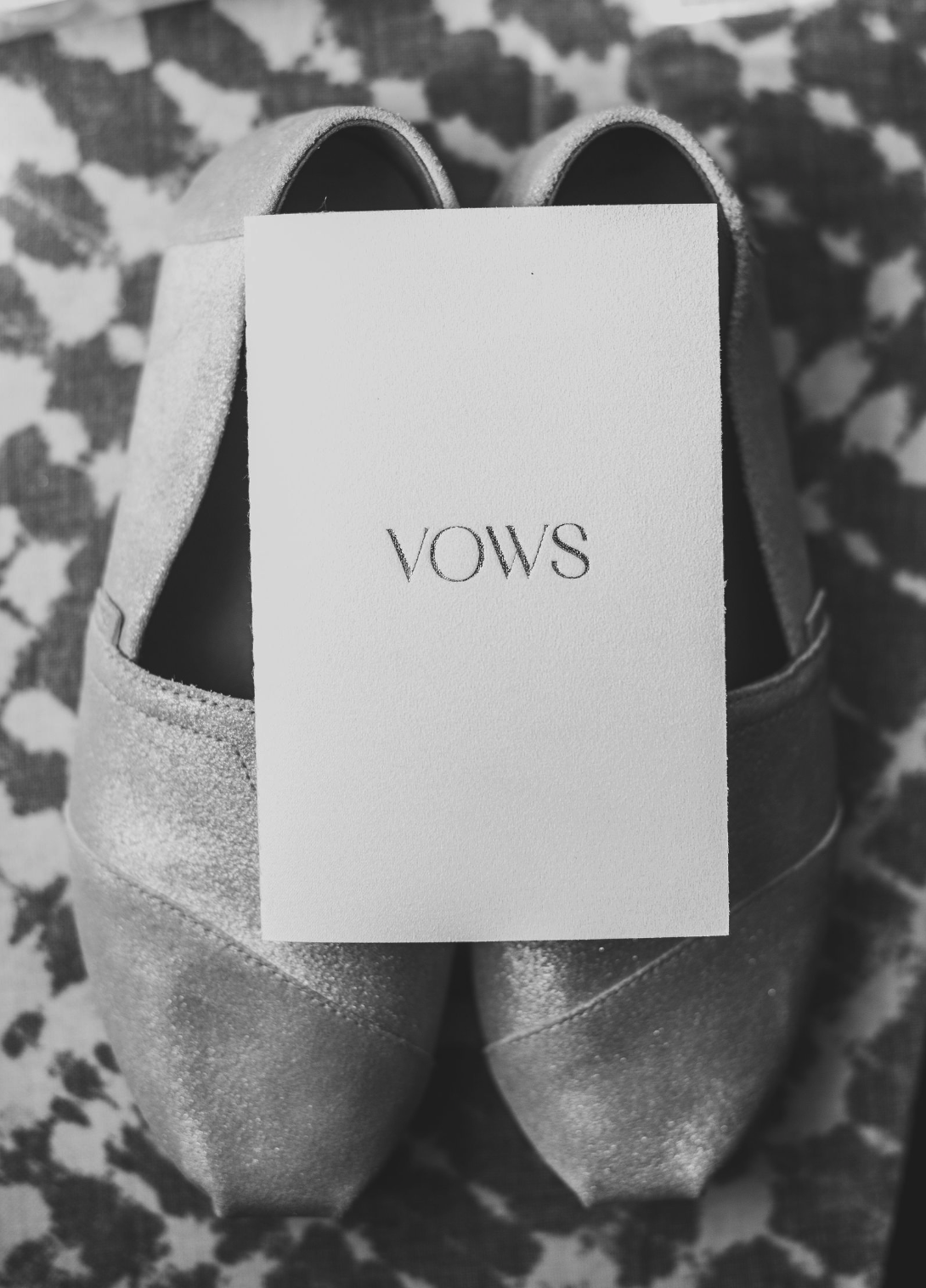 A pair of glittery high heels with a note card labeled 'VOWS' resting on top, on a patterned surface.