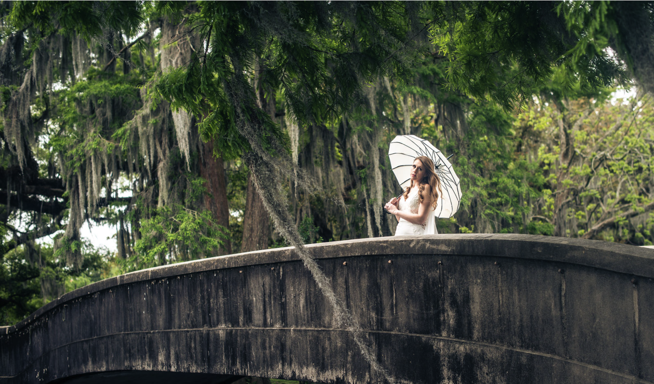 A woman in a white dress holding a white umbrella standing on a curved stone bridge surrounded by green trees with moss hanging from the branches, taken by J. Bellocq, luxury wedding photojournalist of Bellocq Weddings in New Orleans.