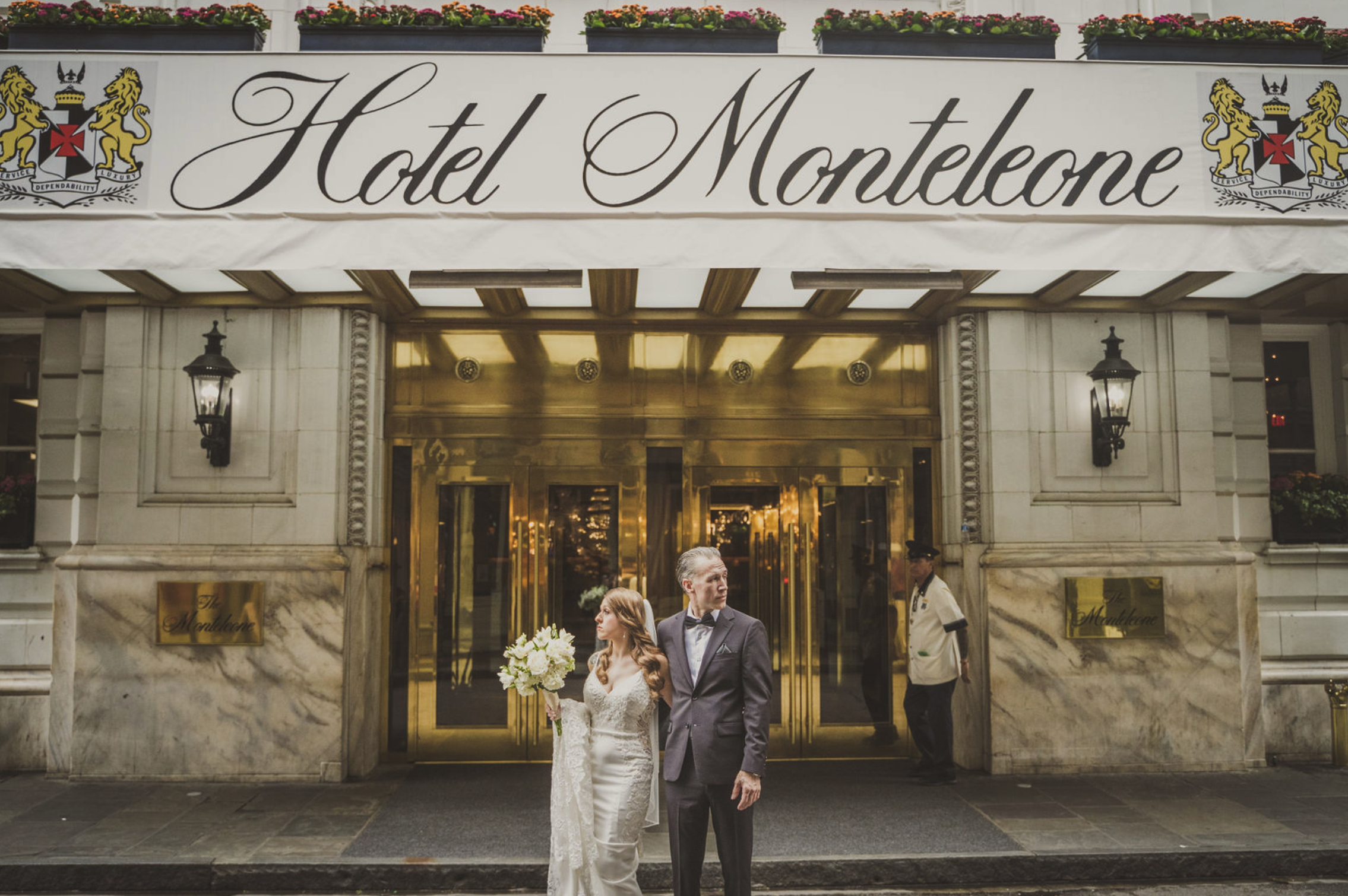 A bride and groom stand outside Hotel Montelone, with the bride holding a bouquet of white flowers. The hotel entrance features golden doors and ornate lanterns, taken by J. Bellocq, luxury wedding photojournalist of Bellocq Weddings in New Orleans.