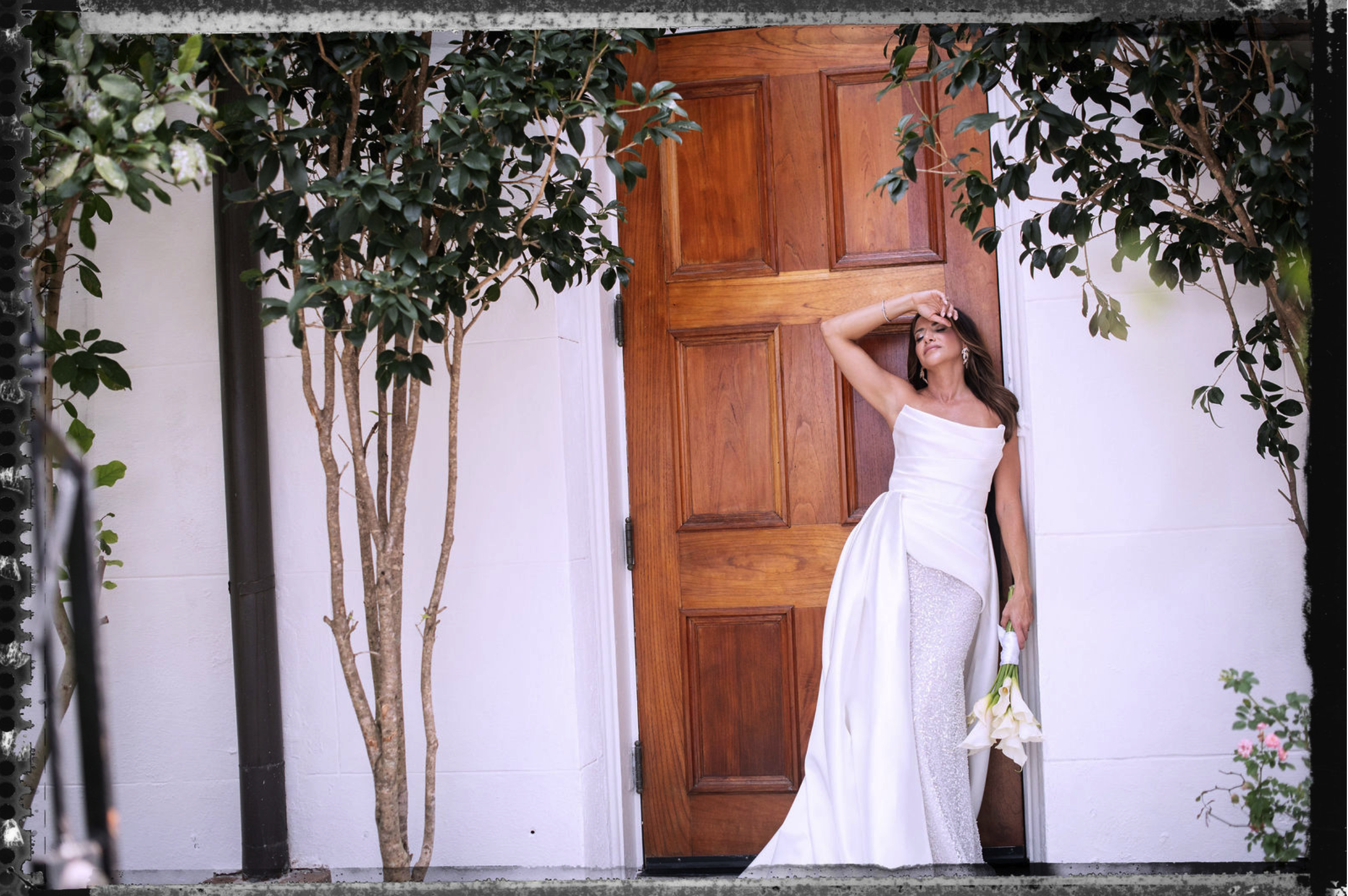 A woman in a white wedding dress stands against a wooden door, holding a bouquet of white flowers. She is touching her forehead and tilting her head back, with her eyes closed. There are two green trees on either side of her, framing the scene.