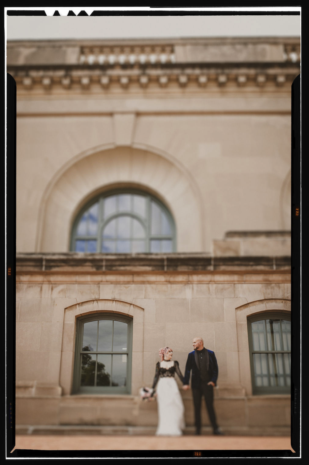 A punk rock, rock n' roll bride and groom holding hands outside a historic stone building with arched windows, taken by J. Bellocq, luxury wedding photojournalist of Bellocq Weddings.