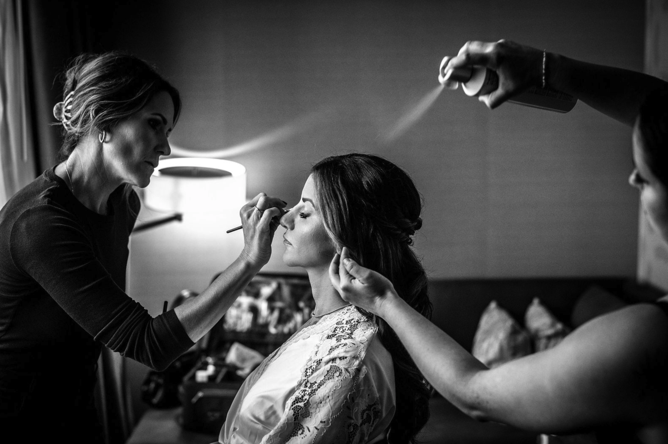 A bride with dark hair is getting her makeup done by a professional makeup artist, while another artist spray curls her hair in a hotel room, black and white photo.