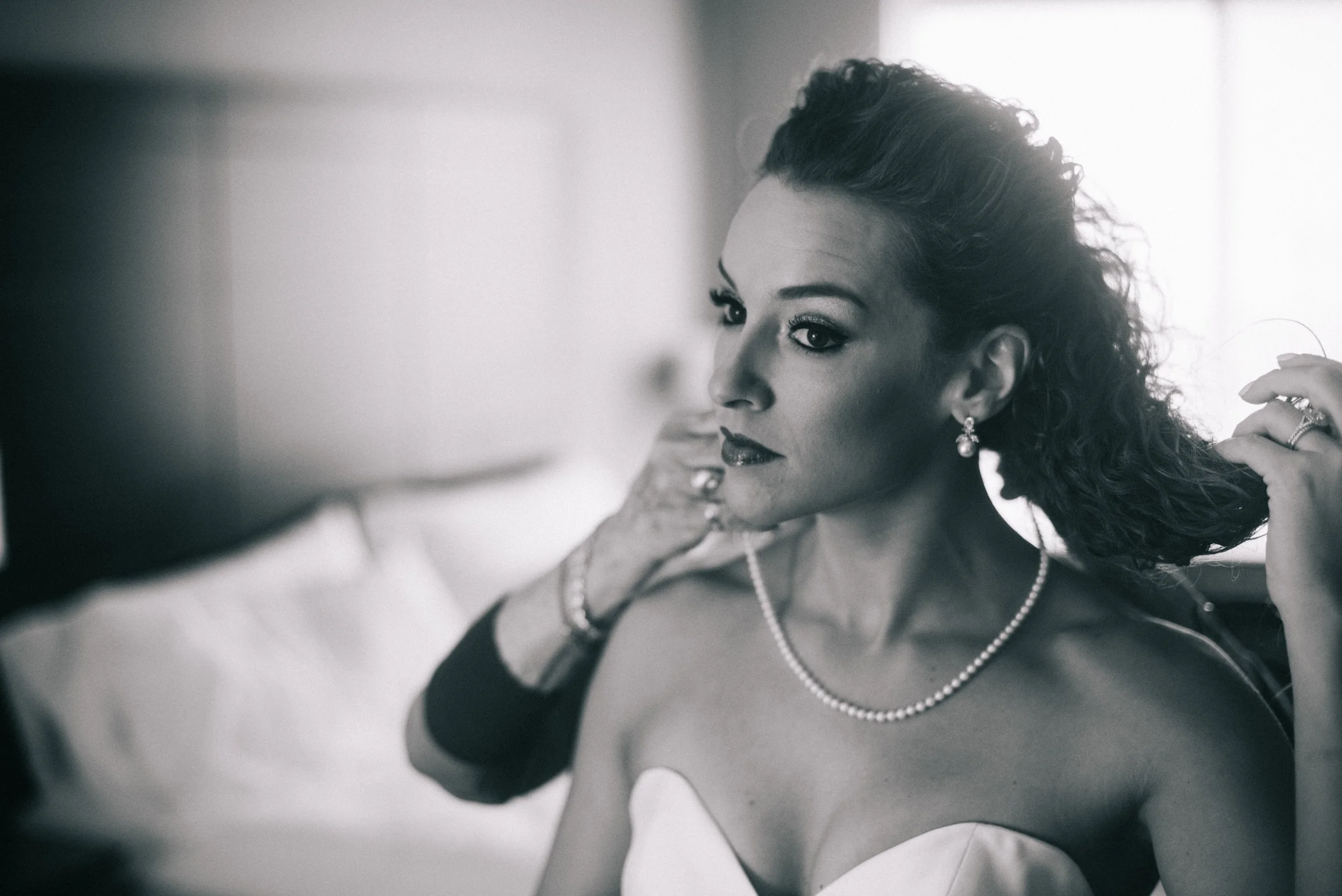 A black and white photo of a woman with dark, curly hair styled in an updo, wearing earrings, a pearl necklace, and a strapless dress, looking directly at the camera while adjusting her hairstyle.