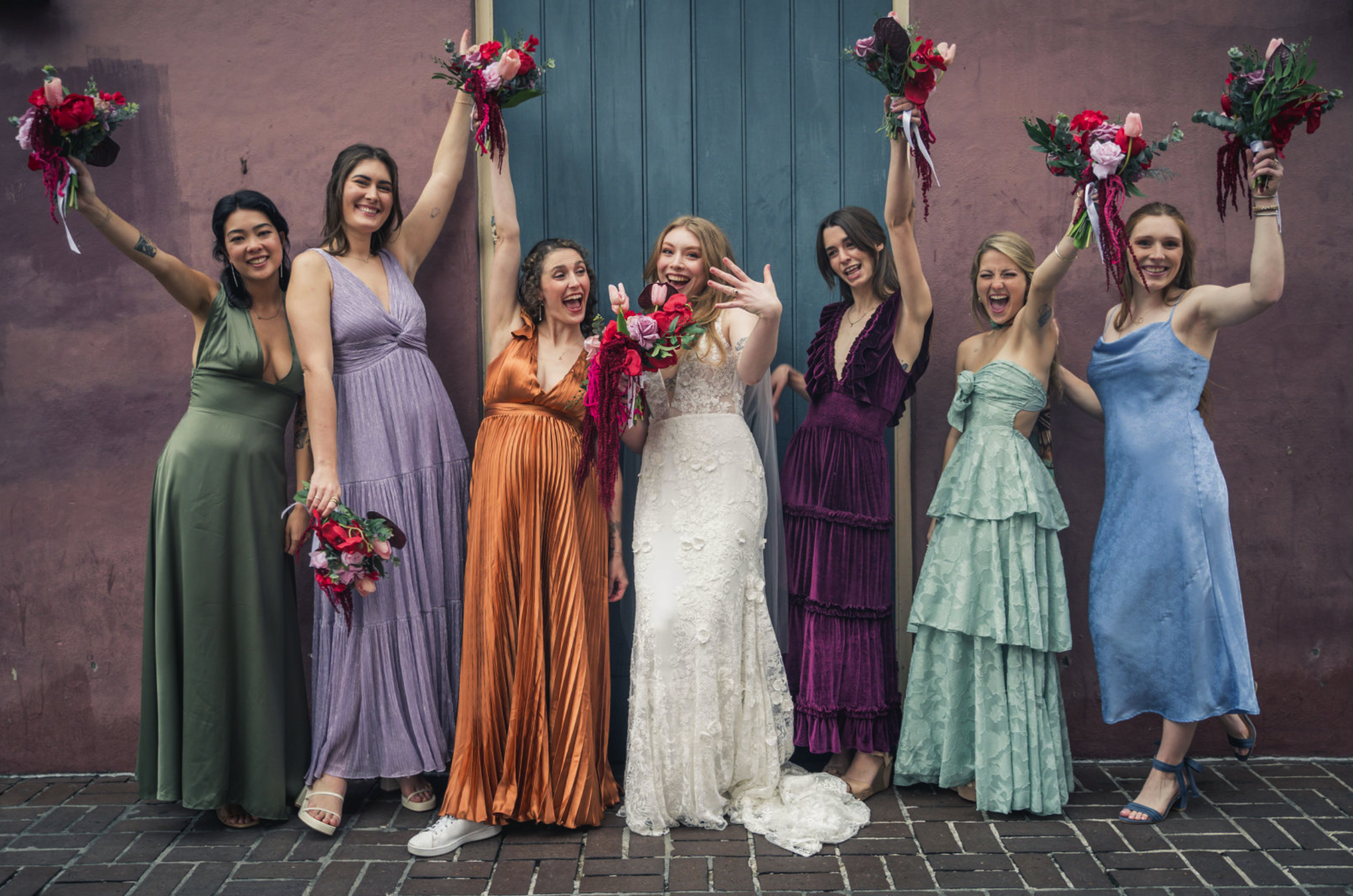 A group of bridesmaids in colorful dresses celebrating a wedding, holding bouquets and raising their arms in joy, standing in front of a brick wall with a blue door, taken by J. Bellocq, luxury wedding photojournalist of Bellocq Weddings