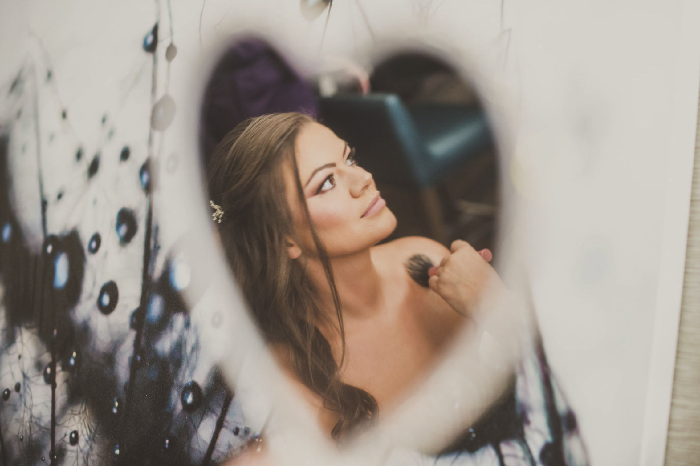 A woman looks at her reflection in a decorative mirror while getting ready, with her hand holding a makeup brush near her shoulder.