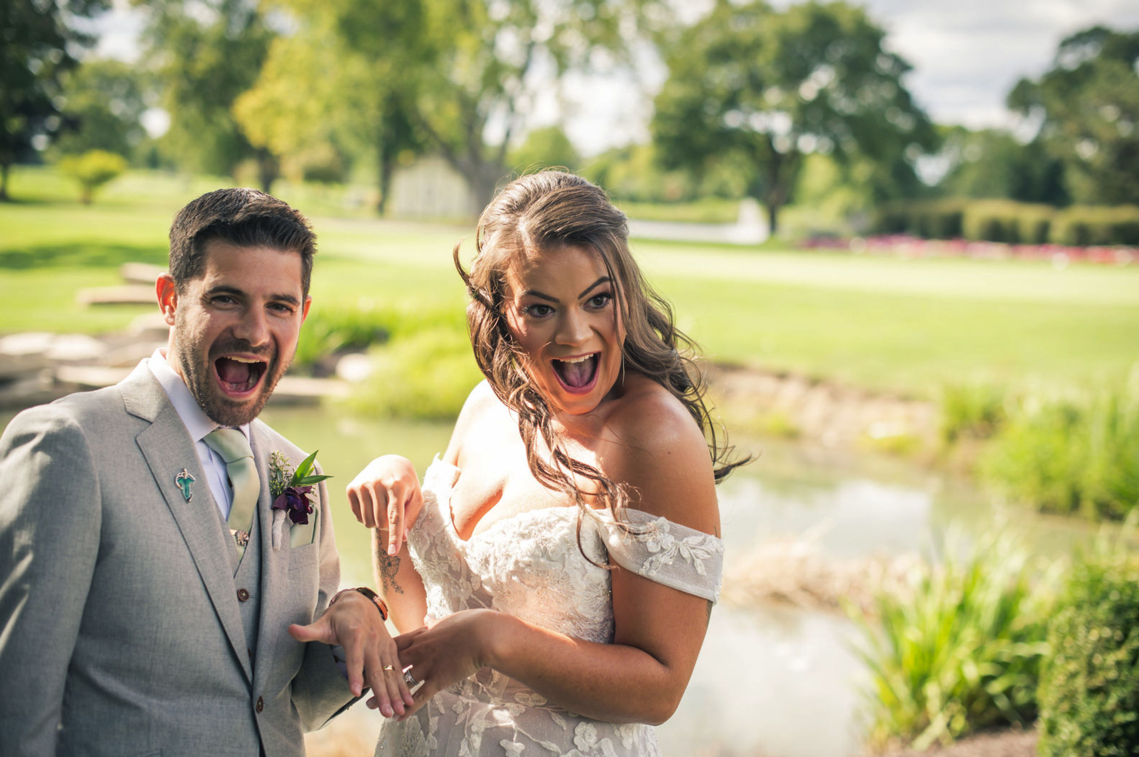 A happy couple in wedding attire celebrating outdoors by a small river in a park, with lush green grass and trees in the background.