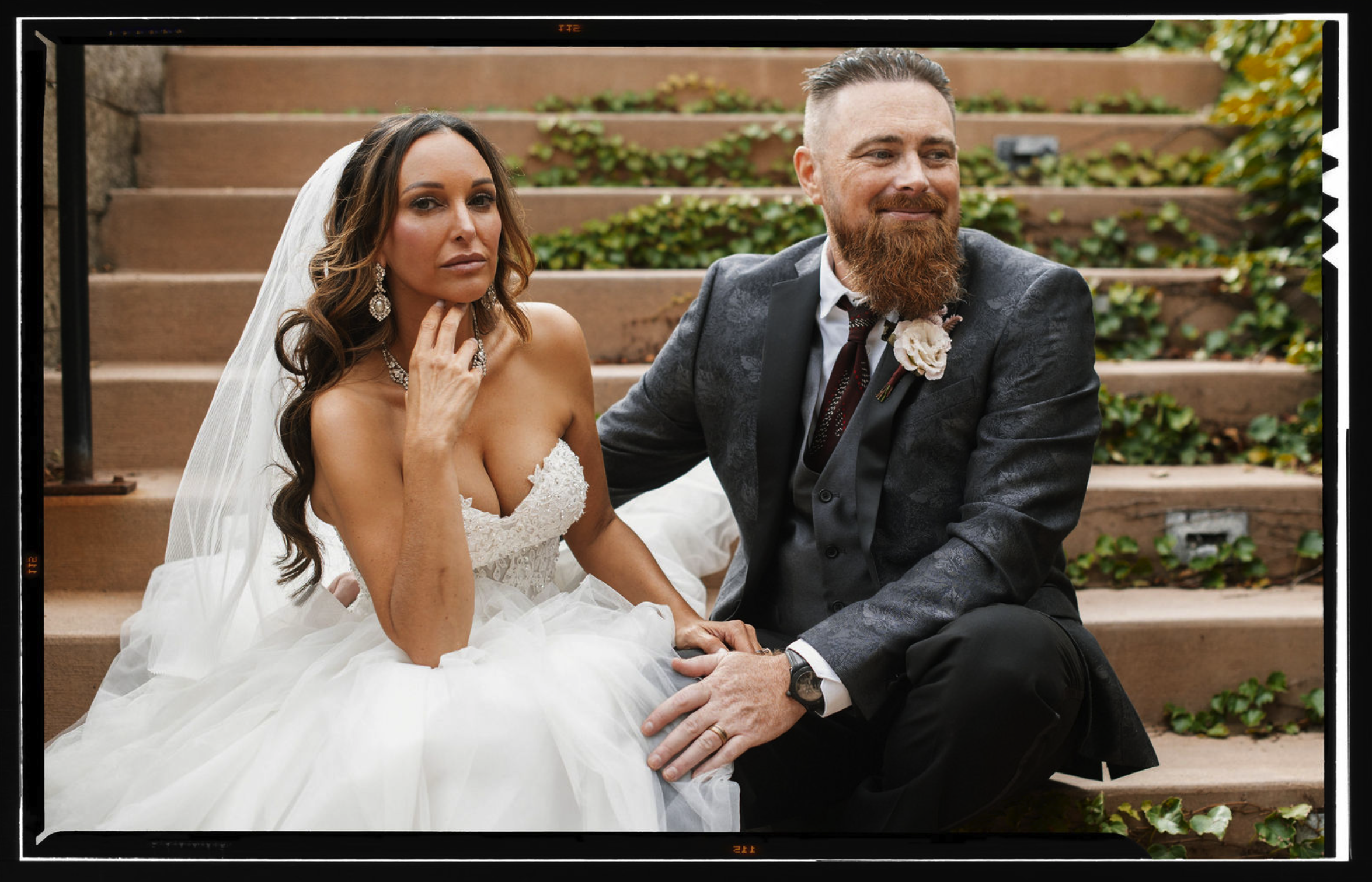 A bride and groom sitting on outdoor steps surrounded by greenery during their wedding photos.