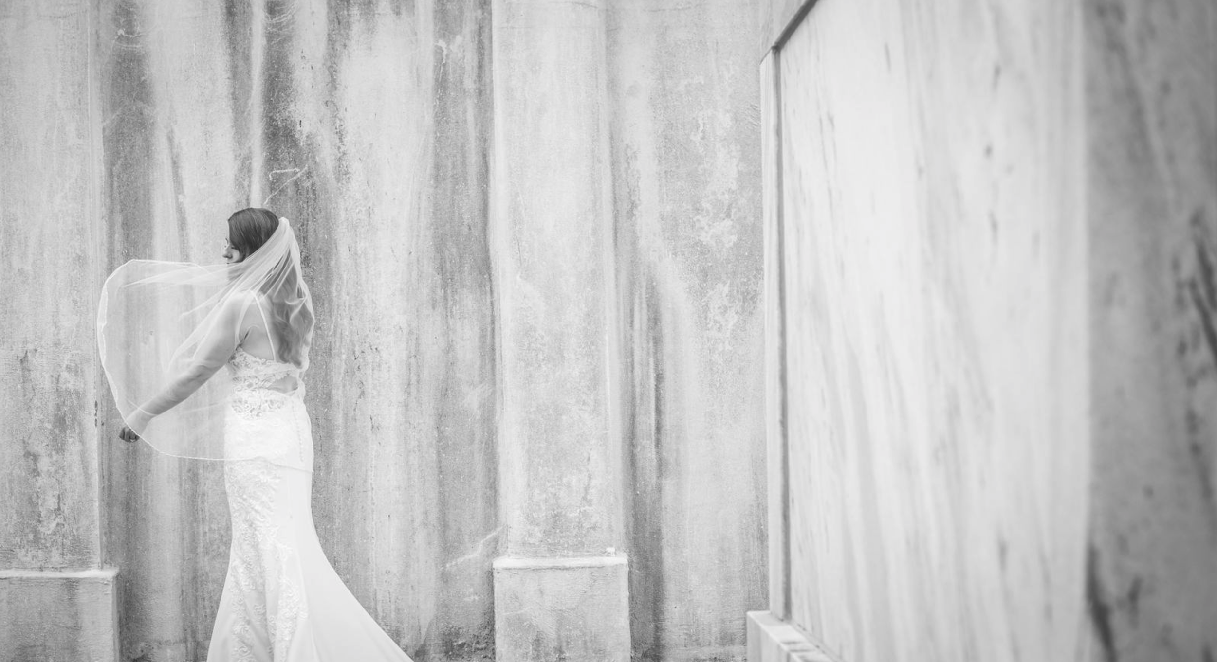 A bride in a wedding dress and veil standing against a textured concrete wall, looking to the left.
